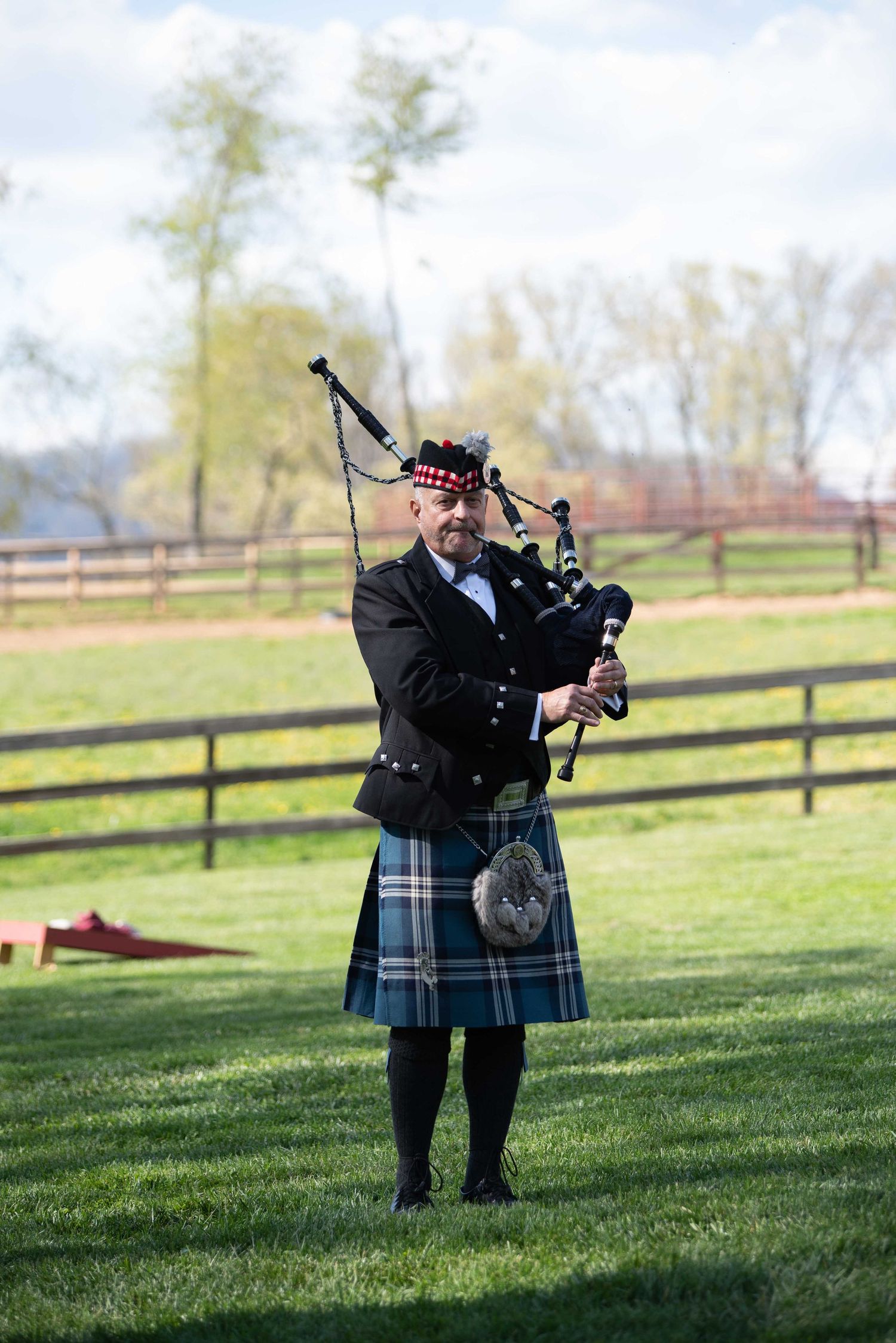 A bagpiper in traditional Scottish tartan kilt and black jacket plays bagpipes on a green lawn at an outdoor event.