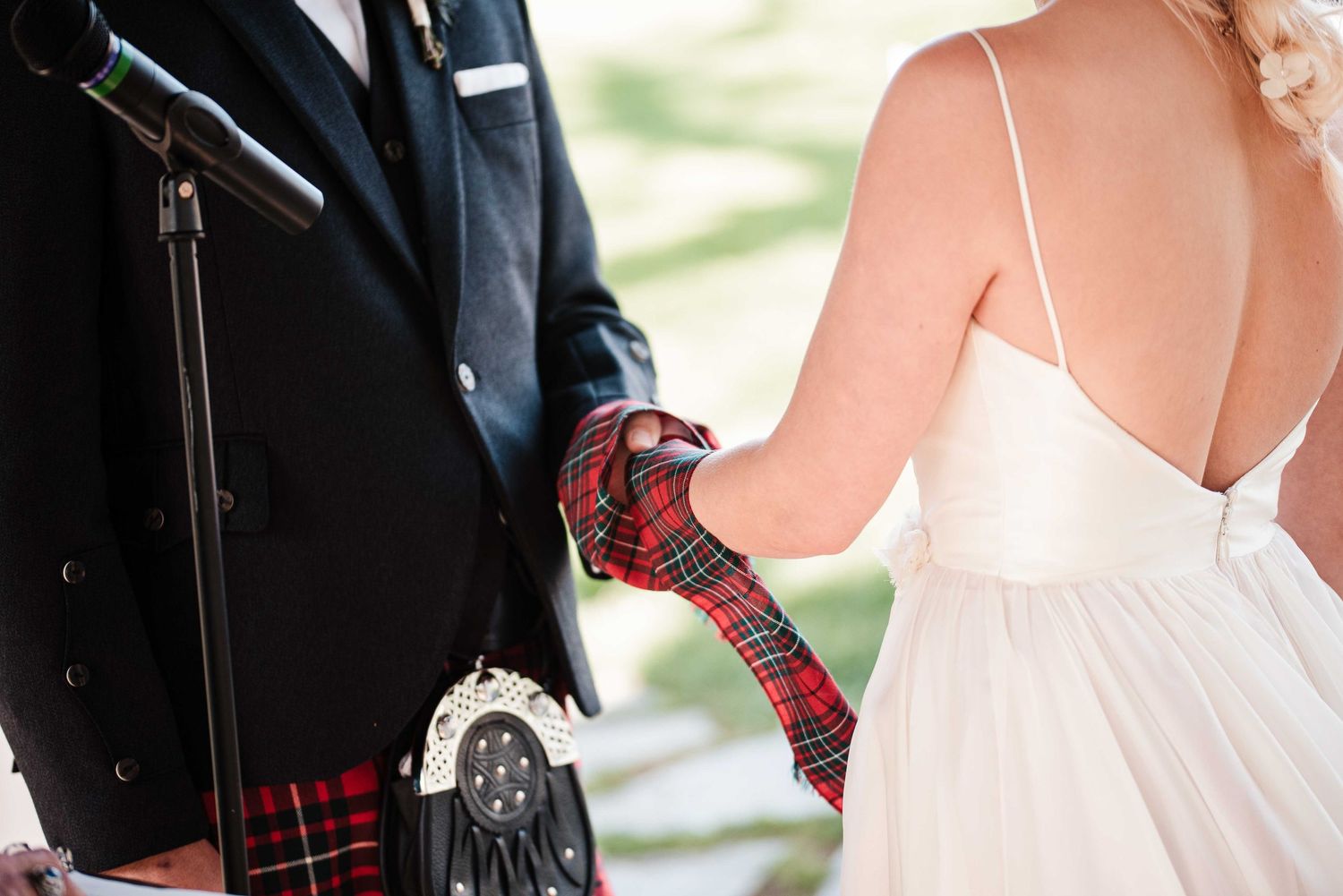 Bride and groom in Scottish kilt with tartan handfasting ribbon during outdoor wedding ceremony.