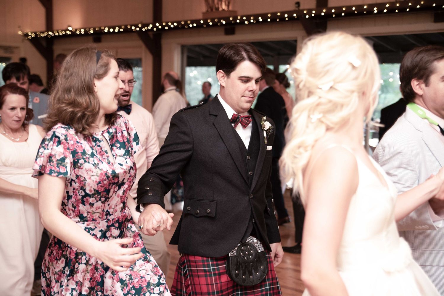 Wedding guests dancing together at a reception, man in red tartan kilt holding hands with woman in floral dress.