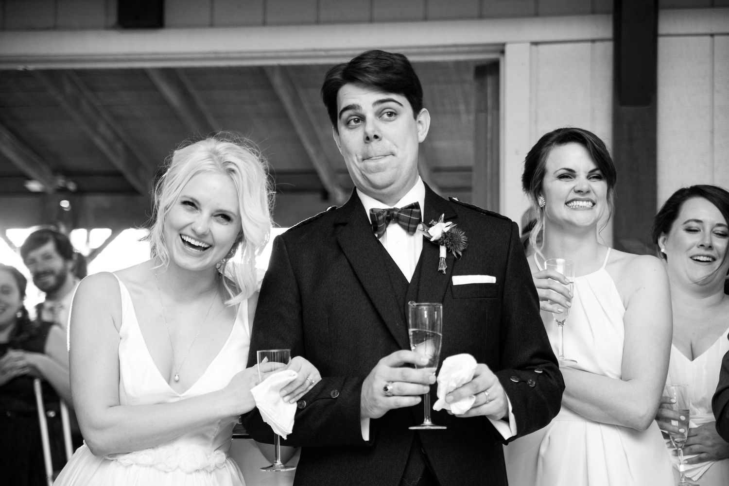 Groom makes funny face holding champagne glass flanked by bridesmaids at wedding reception.