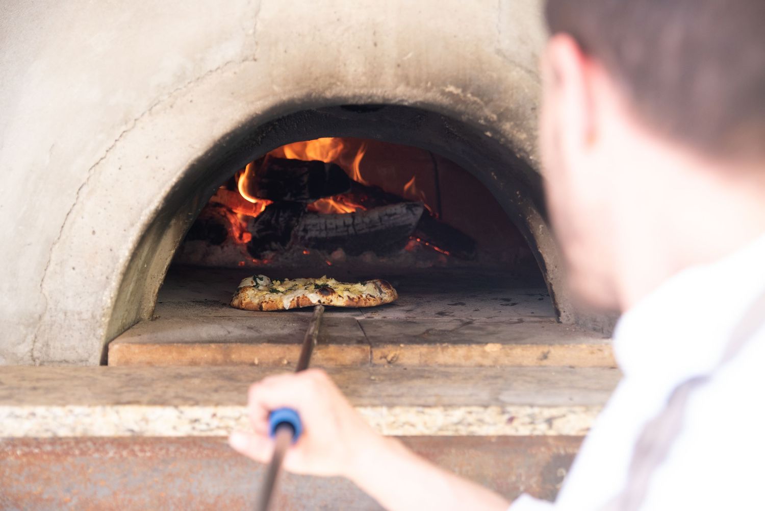 Chef using a peel to cook pizza in a wood-fired stone oven with roaring flames.