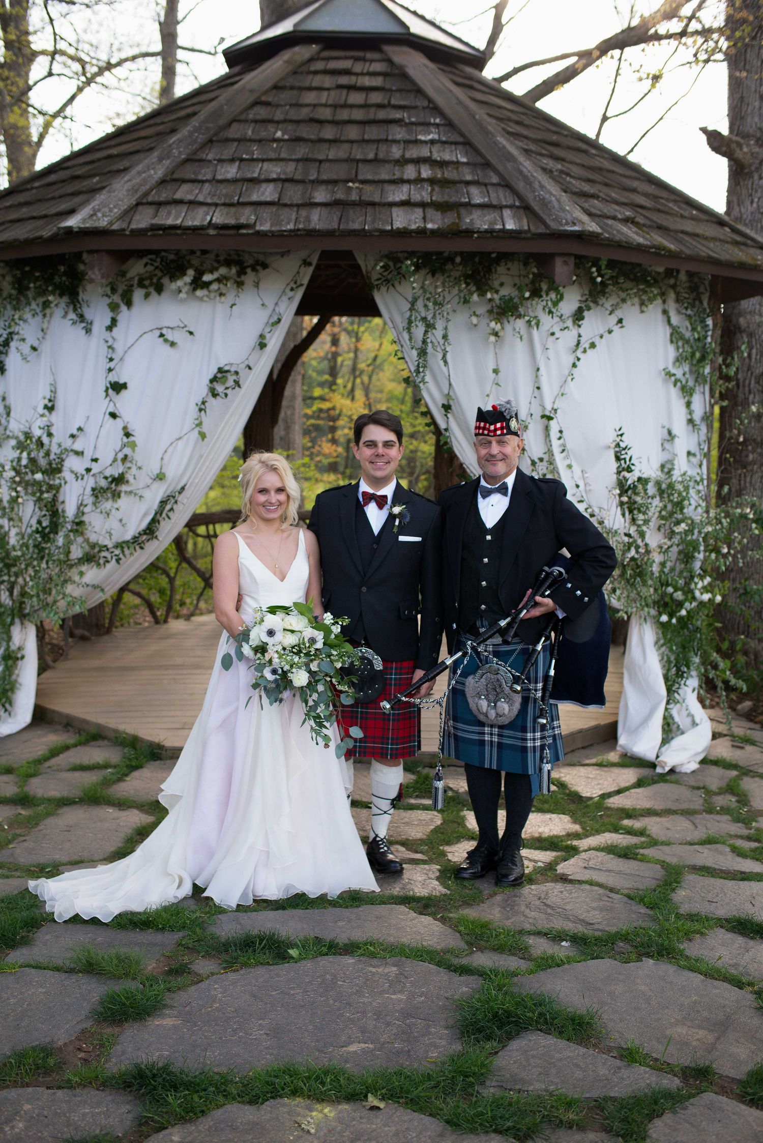 Bride and groom pose with Scottish bagpiper in tartan kilt at outdoor gazebo wedding ceremony.