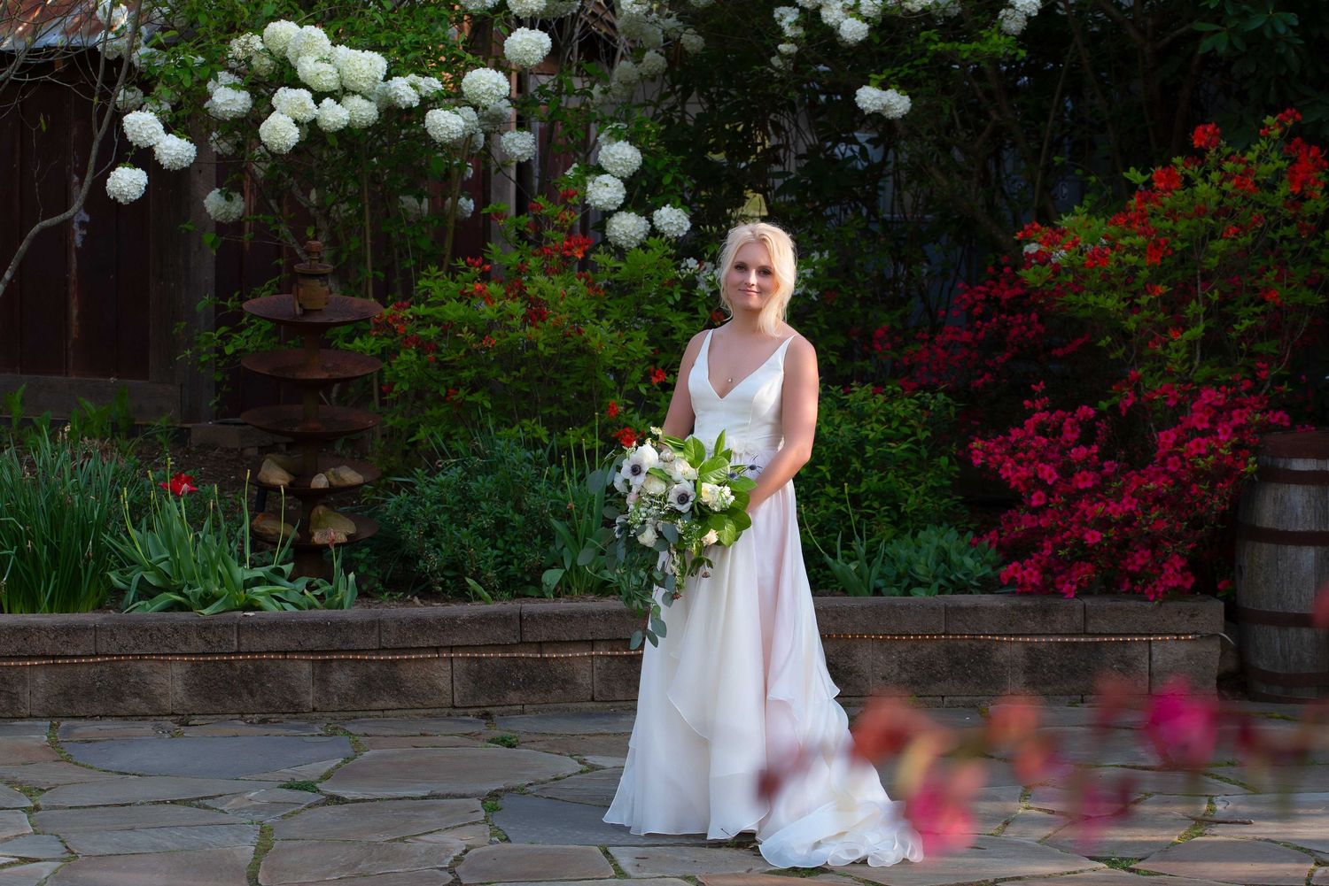 Bride in white gown holding bouquet stands on stone patio surrounded by lush colorful garden blooms.
