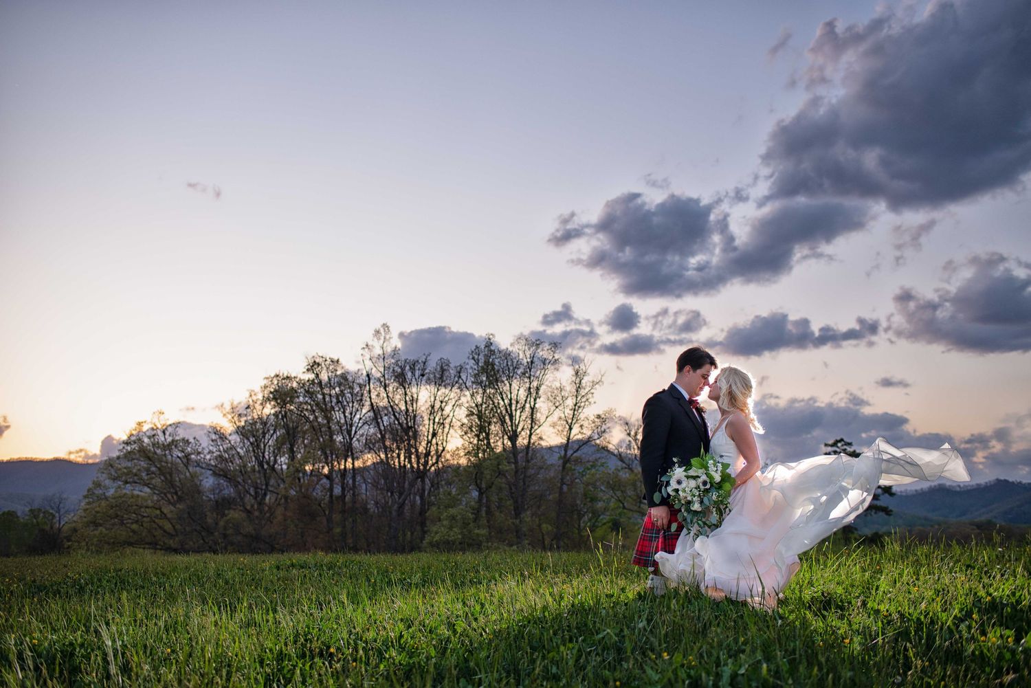 Bride and groom share a romantic kiss in a lush green field at sunset with mountains in the background.