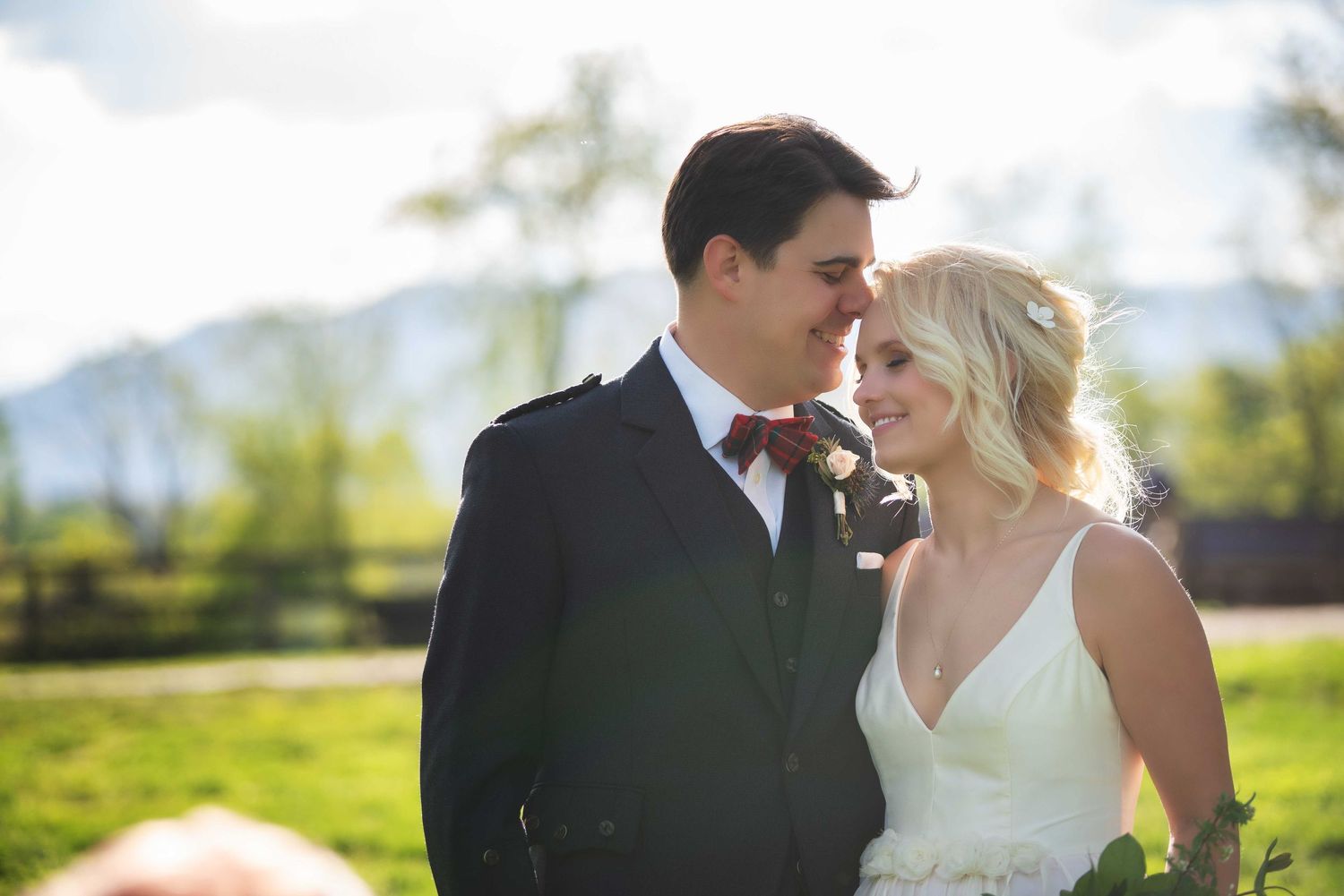A bride and groom share a romantic moment outdoors with green fields and mountains in the background on their wedding day.