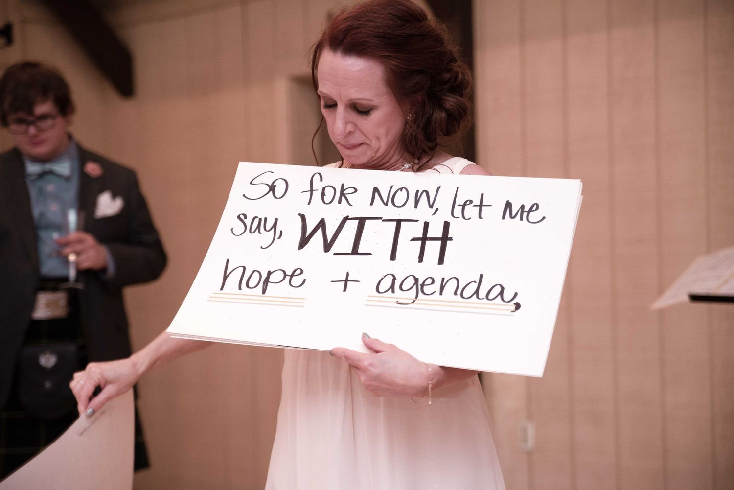 Woman in pink dress holds handwritten sign reading 'so for now let me say, WITH hope + agenda' at formal event.