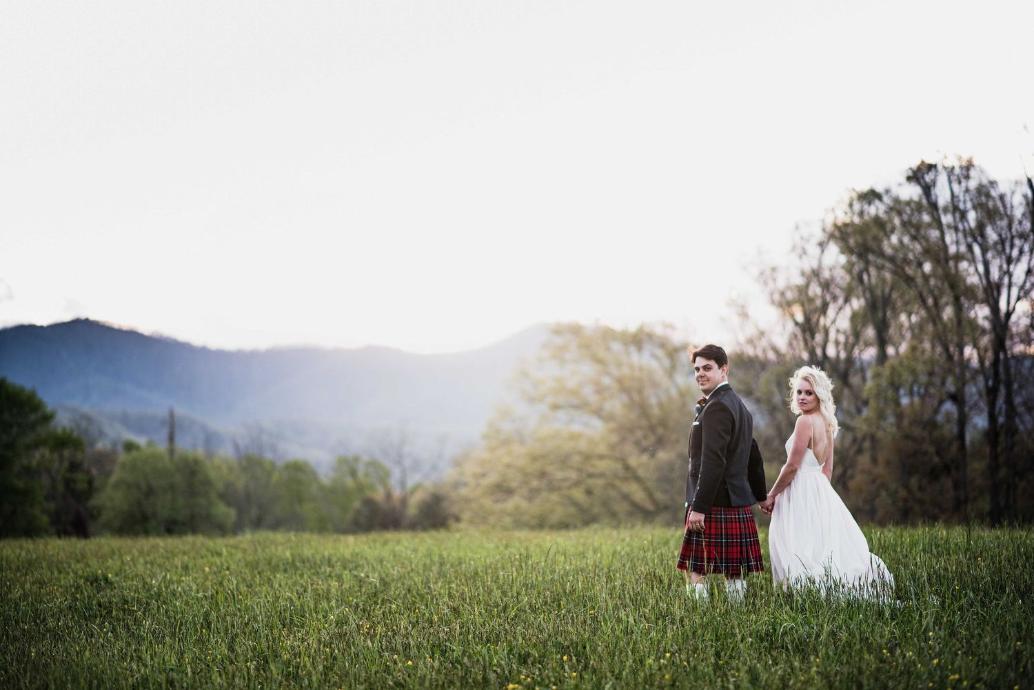 Bride and groom in Scottish kilt stand in lush green meadow with mountain backdrop at golden hour.