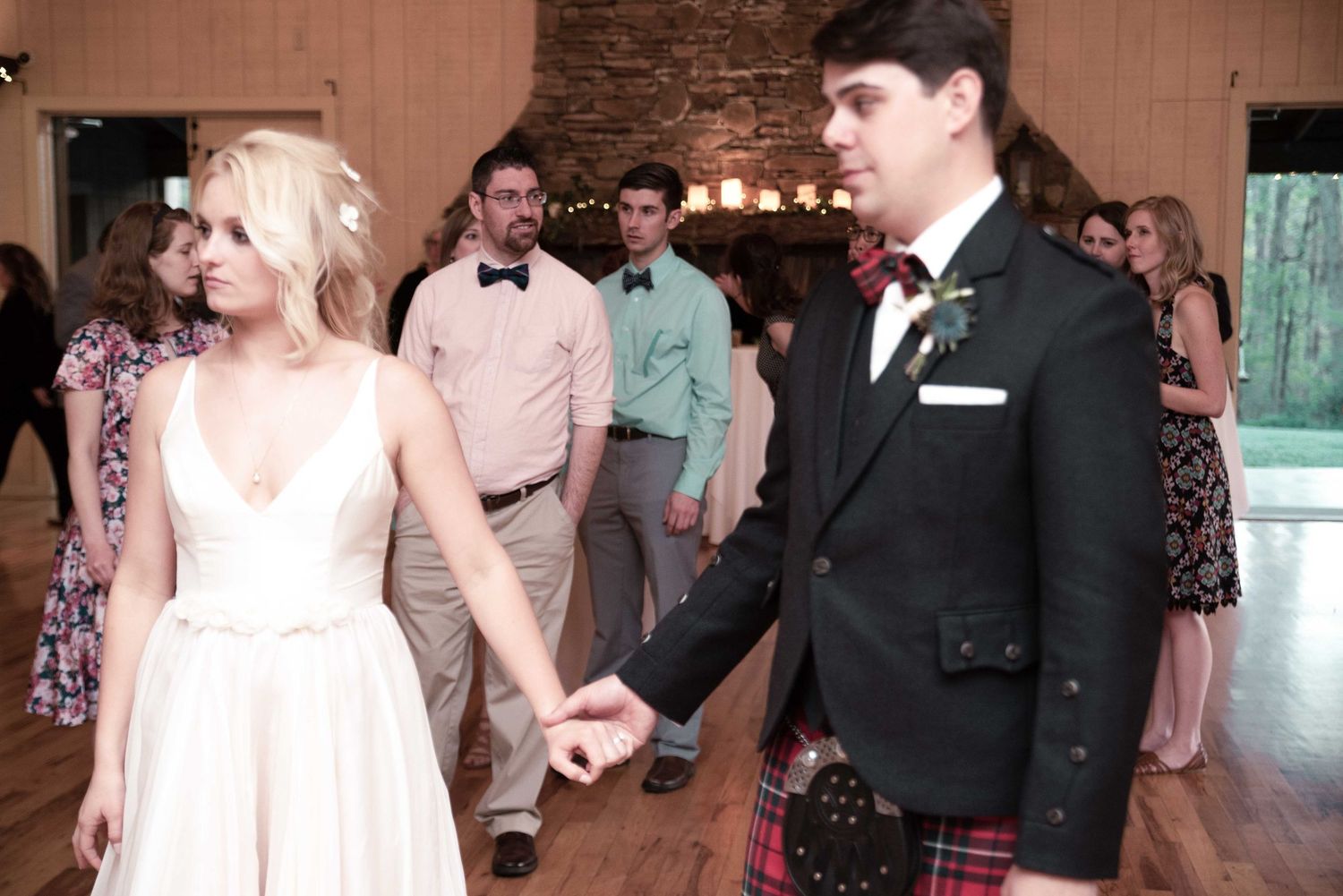 Bride in white gown and groom in tartan kilt share first dance at elegant rustic wedding reception.