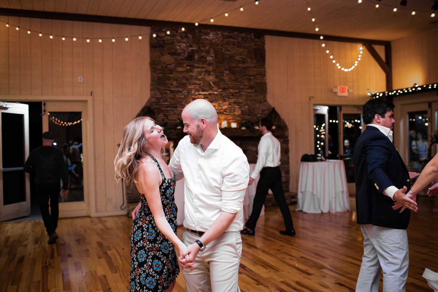 Couple dancing and holding hands at a rustic wedding reception with string lights and stone fireplace.
