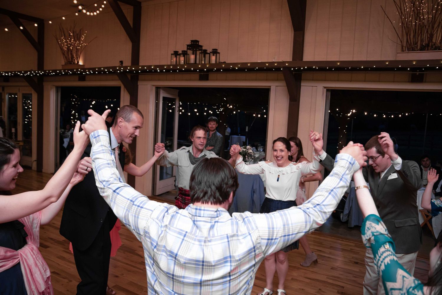 Wedding guests joyfully dancing in a circle at a rustic barn reception with string lights.