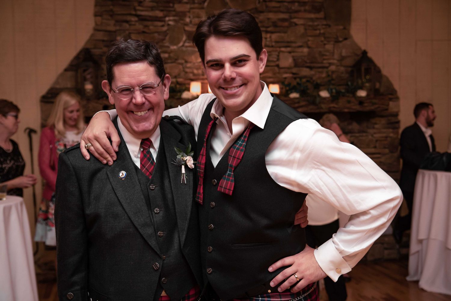 Two men in Scottish kilts and vests smile together at a wedding reception with a stone fireplace backdrop.