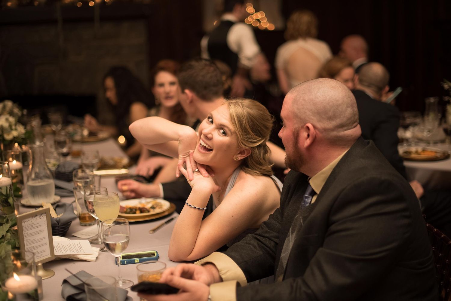 A woman in an elegant black dress smiles warmly at a man during a formal dinner event at a dimly lit venue.
