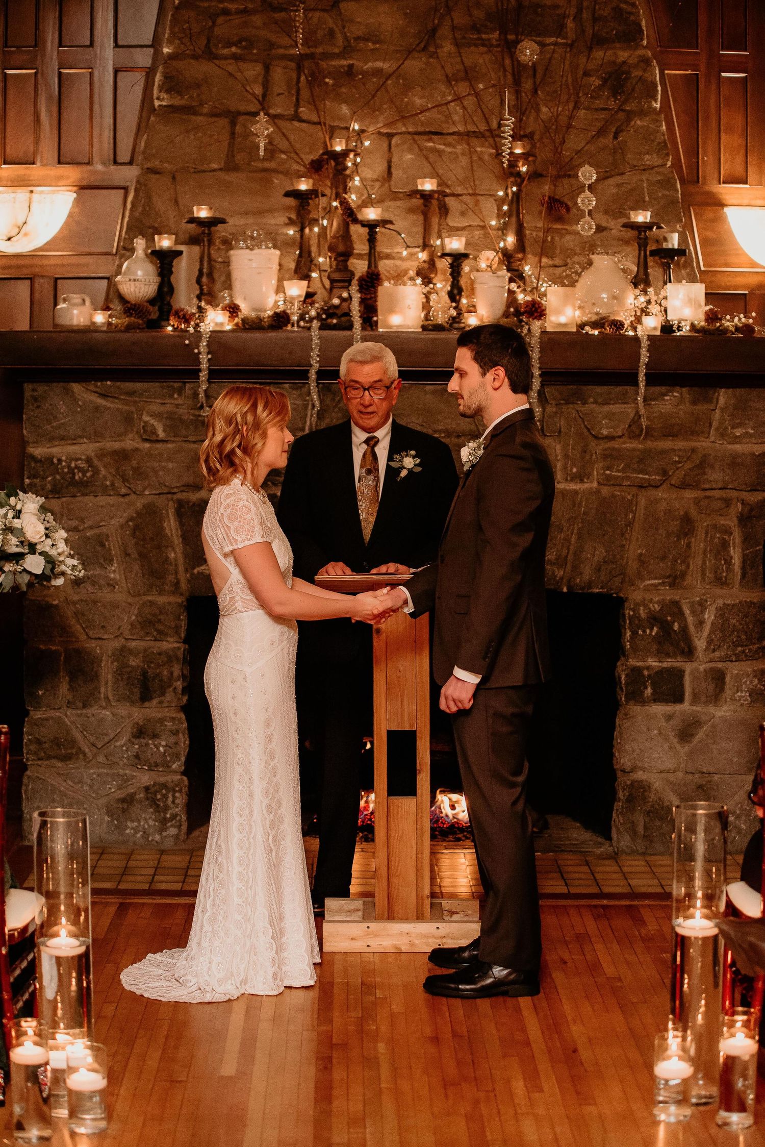 Bride and groom exchange vows at candlelit stone fireplace altar during intimate indoor wedding ceremony.