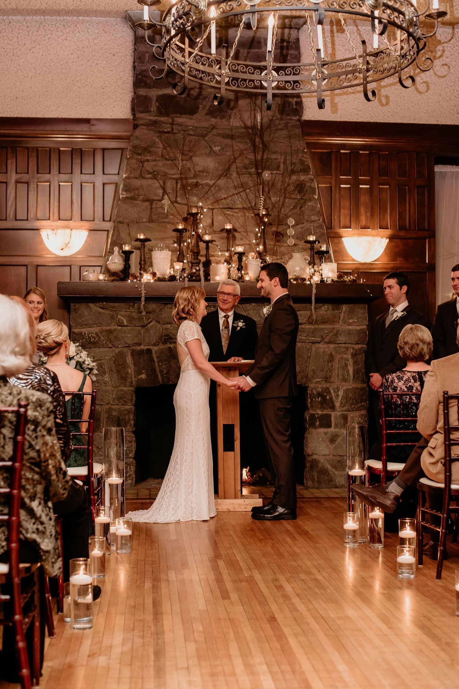 Bride and groom exchange vows before a stone fireplace adorned with candles at an elegant indoor wedding ceremony.