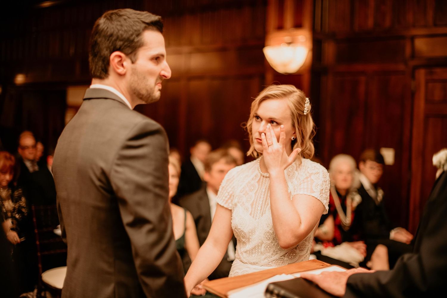 Emotional bride wiping tears during wedding ceremony in elegant wood-paneled venue with guests seated behind.