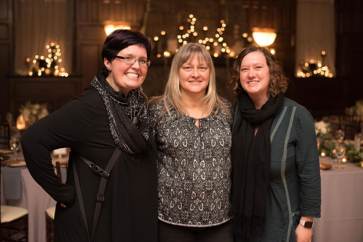 Three women smiling together at an elegant indoor event with warm string lights and decorated tables in the background.