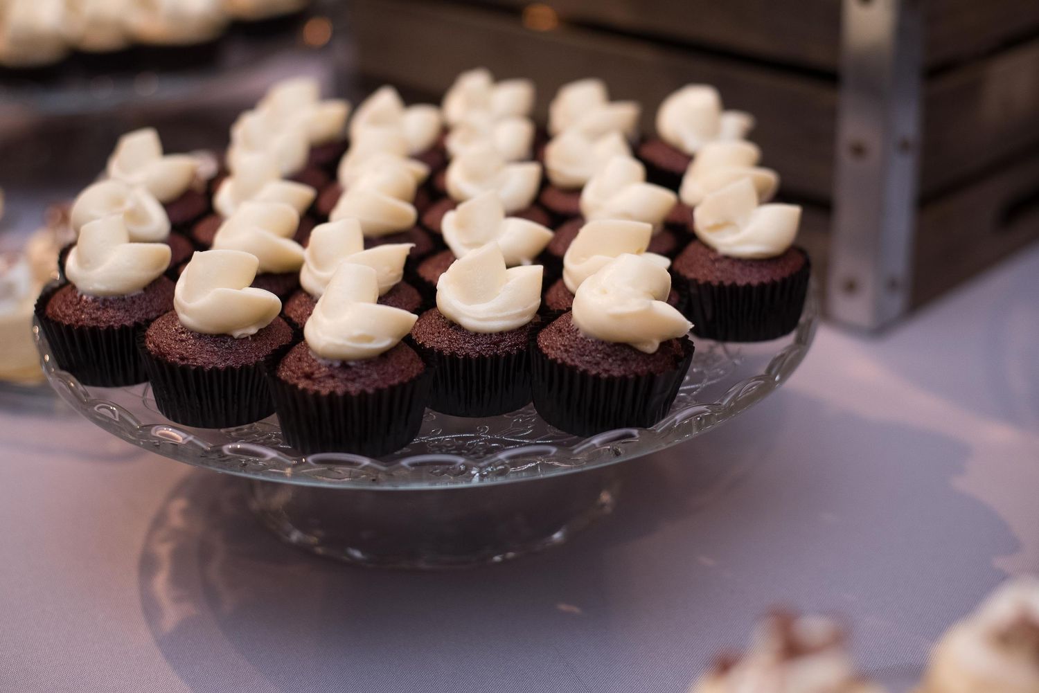 Mini chocolate cupcakes with white cream cheese frosting displayed on a glass cake stand at a dessert table.