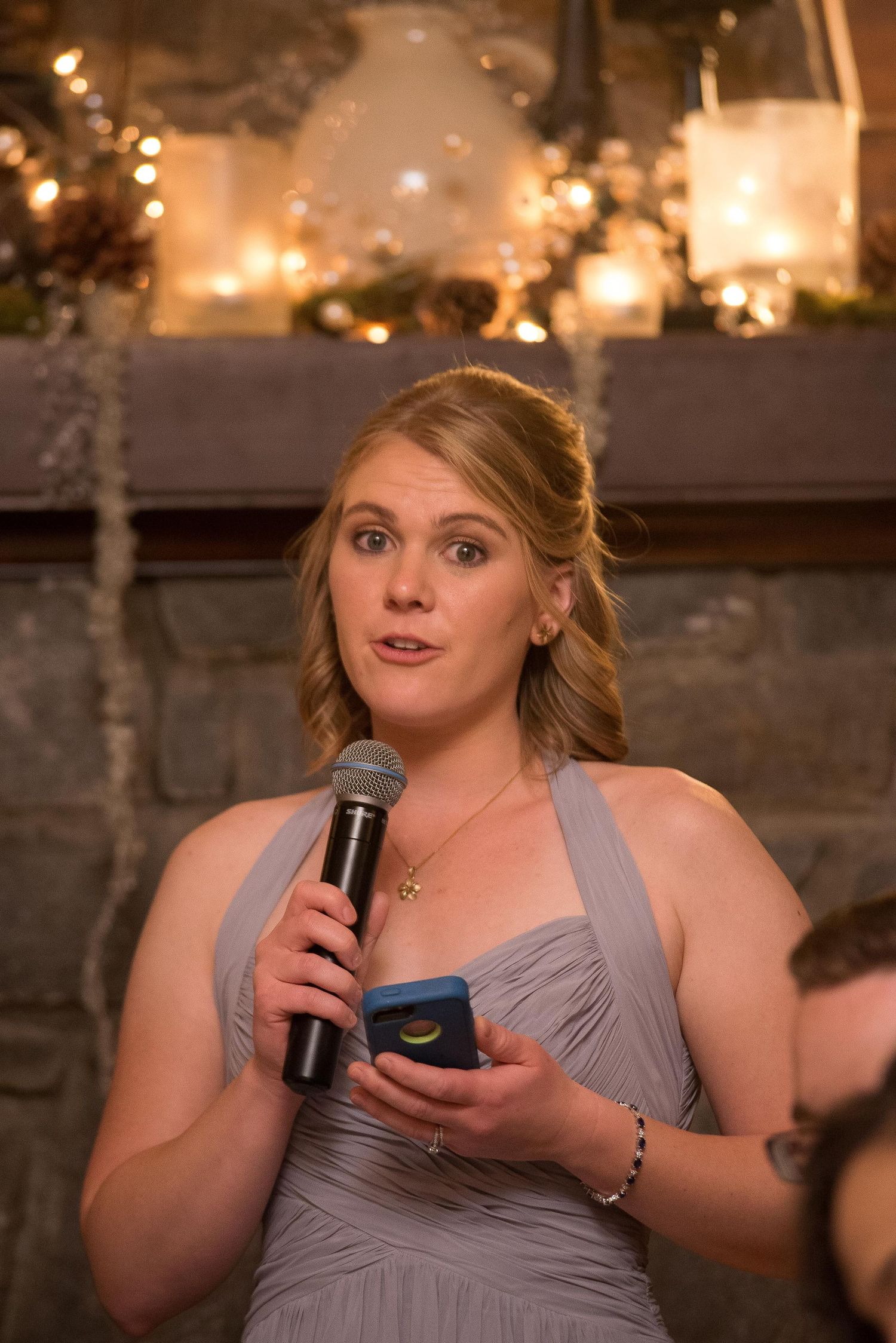 Bridesmaid in gray dress giving a speech at a wedding reception, holding a microphone and notes.