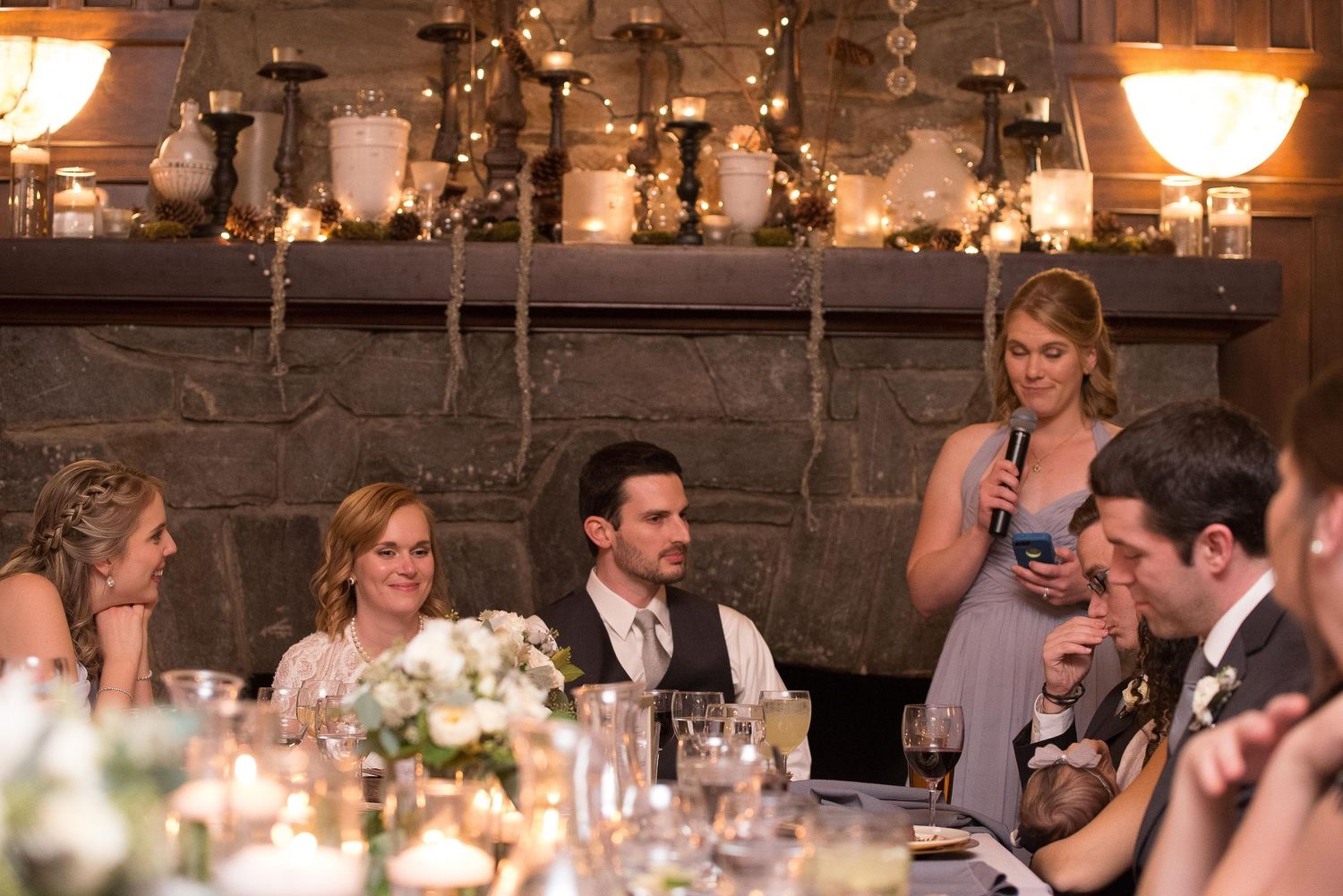 Wedding reception guests gathered around a candlelit table near a stone fireplace, as a bridesmaid delivers a heartfelt toast.