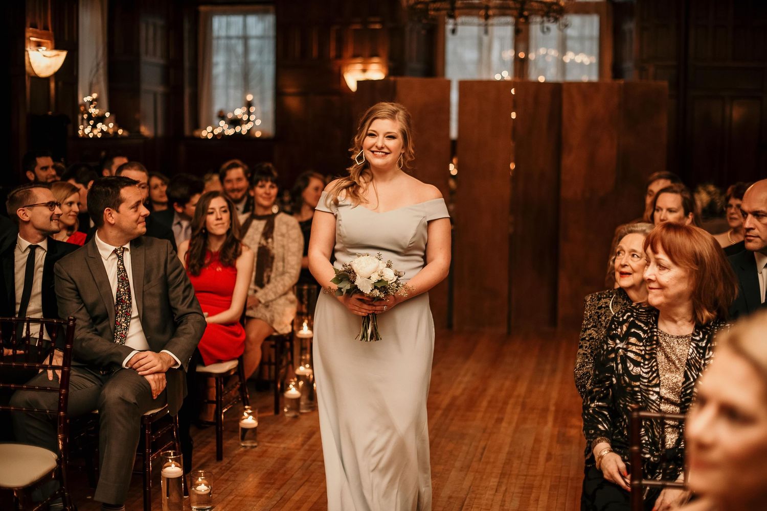 Bridesmaid in silver gown walks down wooden aisle holding white bouquet at elegant indoor wedding ceremony.