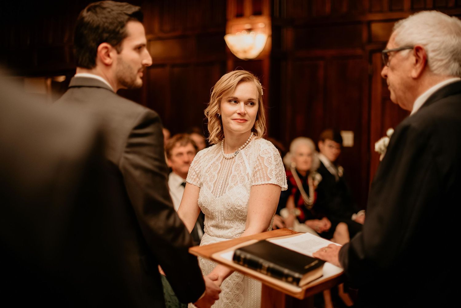Bride smiling at groom during indoor wedding ceremony, officiant holding Bible, guests seated in background.