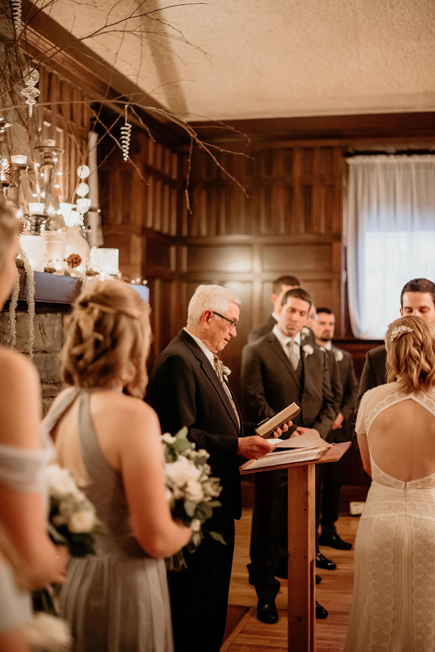 Officiant reads vows at elegant indoor winter wedding ceremony with bridesmaids in gray dresses holding white bouquets.