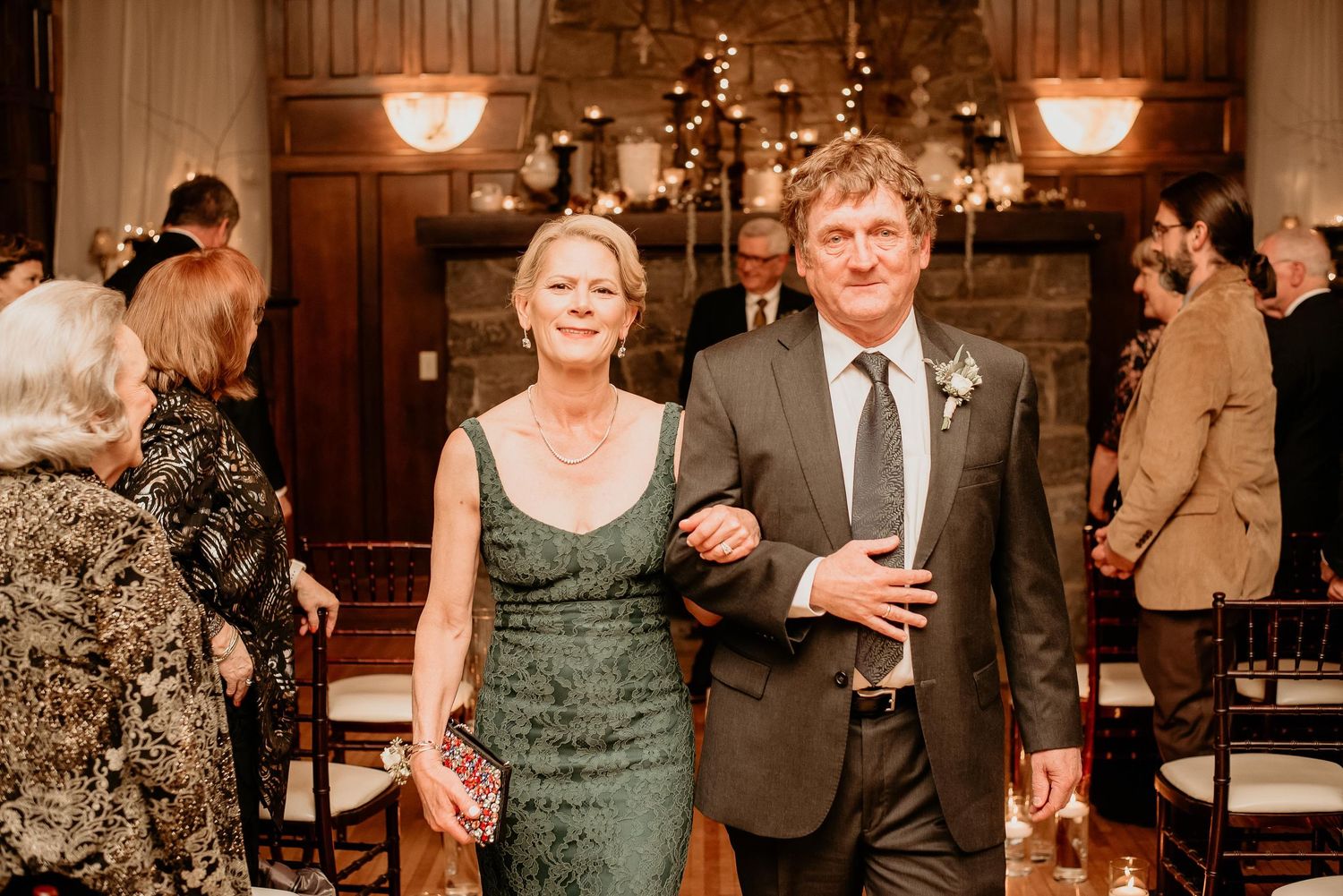 Elegant couple walking arm in arm at a wedding ceremony, woman in green lace dress, man in gray suit with boutonniere.