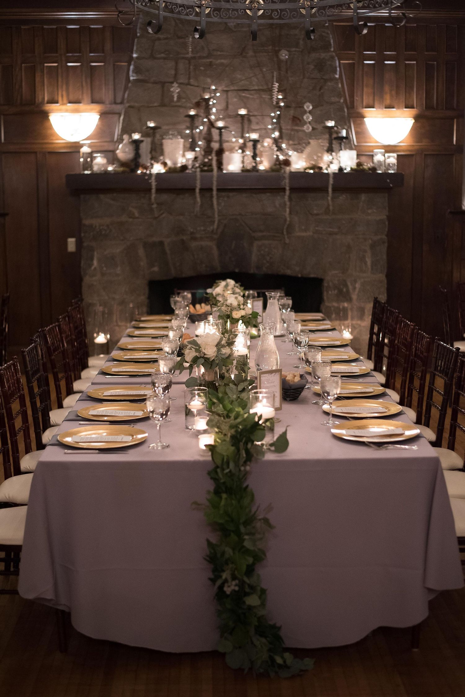 Elegant wedding reception table with greenery runner, gold plates, candles, and stone fireplace backdrop.