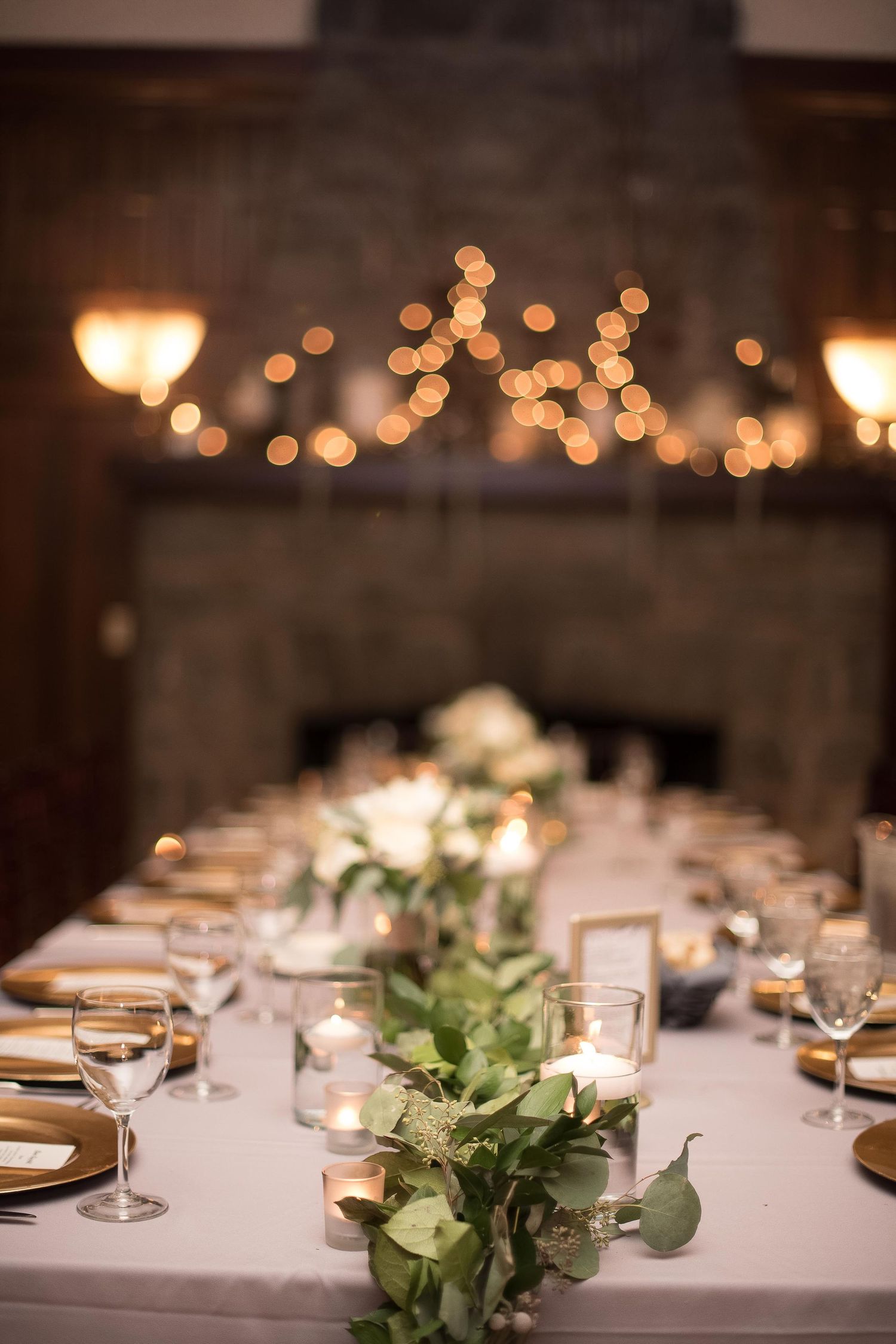 Elegant wedding reception table with greenery runner, white roses, candles, gold chargers, and bokeh string lights near a stone fireplace.