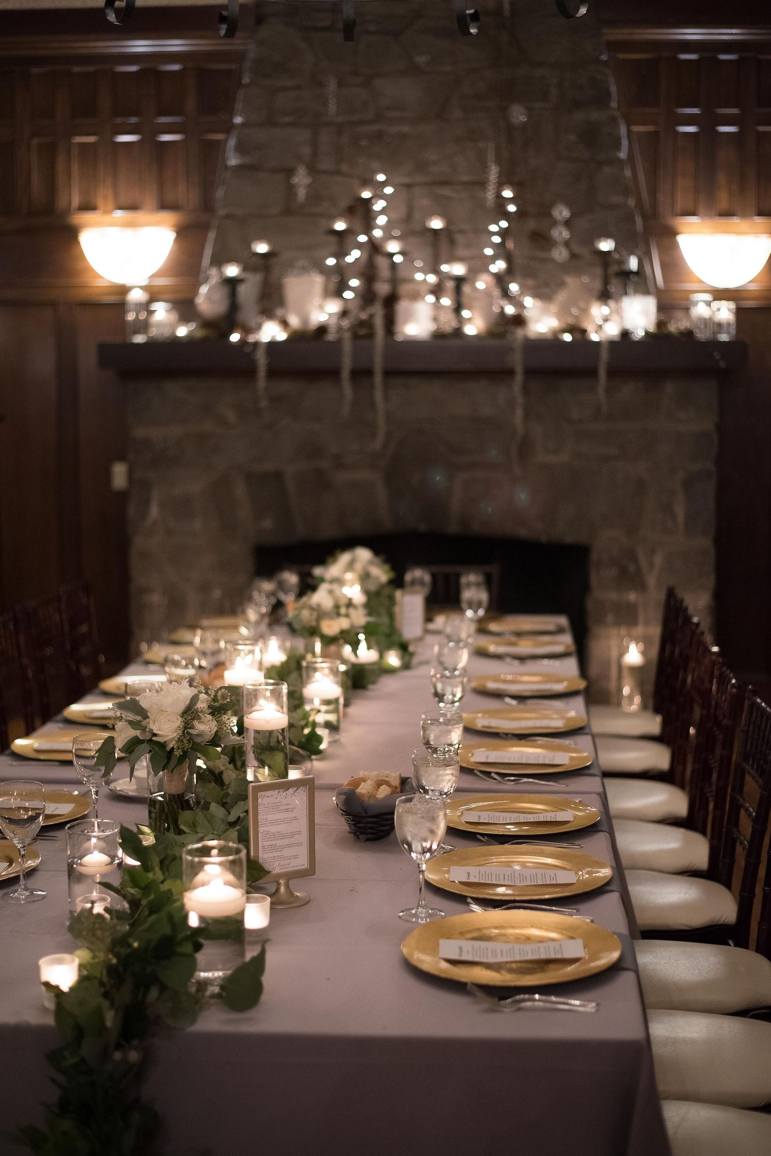 Elegant wedding reception table with gold chargers, greenery runner, candles, and stone fireplace backdrop.