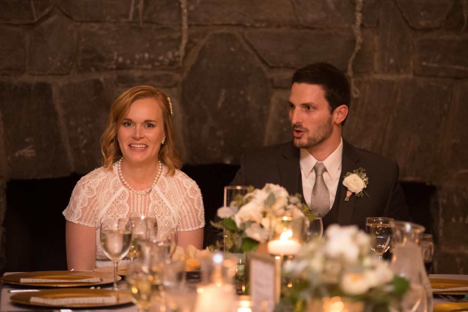 Couple in formal attire seated at elegantly set wedding reception table with candles and floral centerpiece.