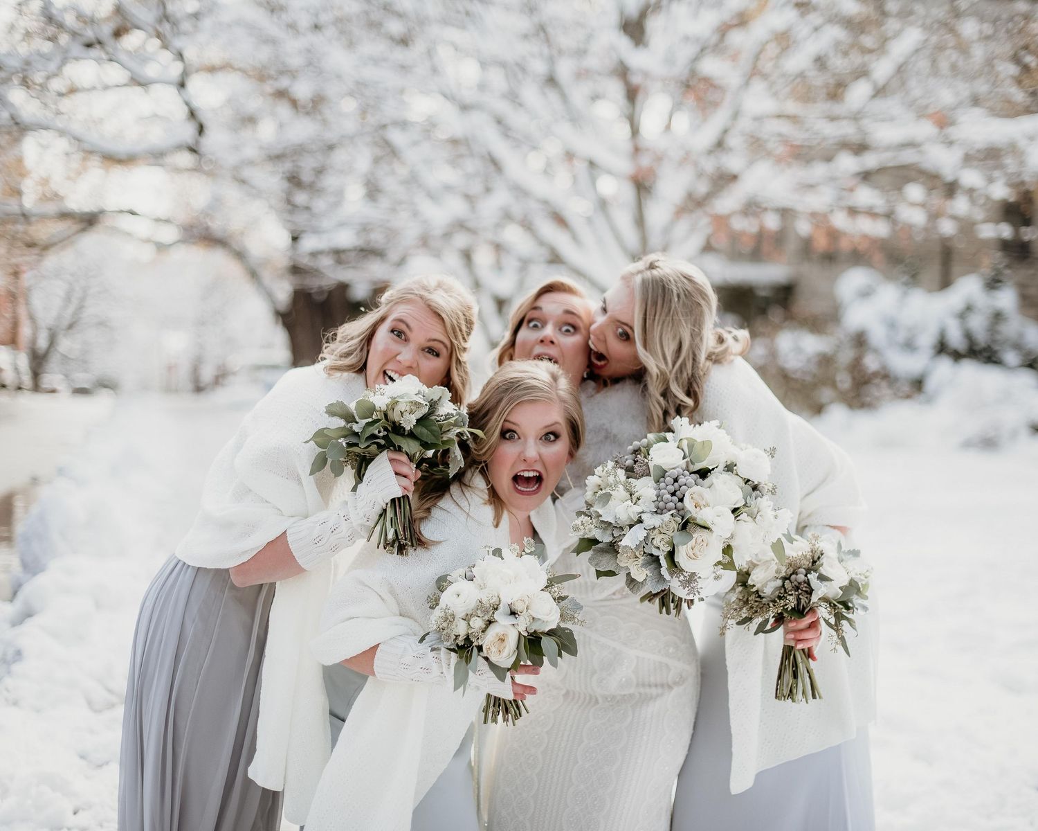 Bride and bridesmaids in white wraps holding bouquets laughing together in a snowy winter wedding outdoor setting.