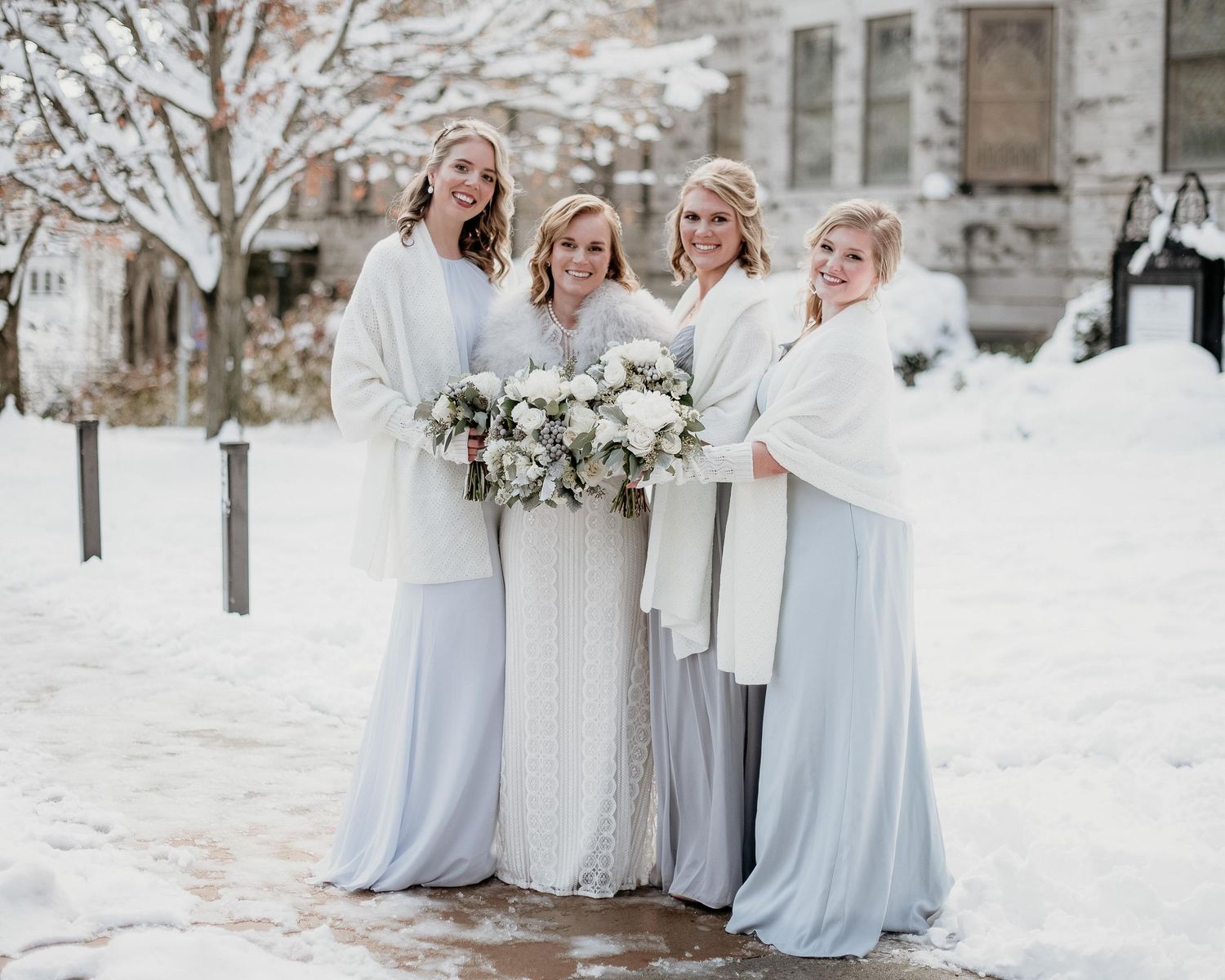 Bride and bridesmaids in white and light blue gowns with fur wraps holding bouquets outdoors in a snowy winter setting.