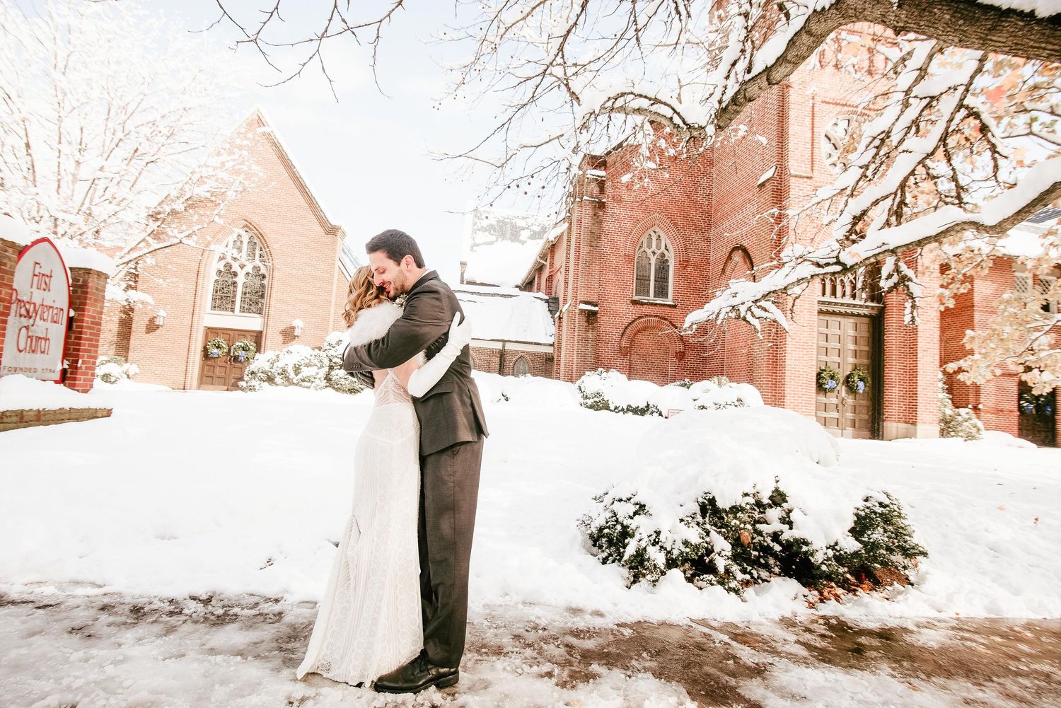 Bride and groom embrace in snow outside red brick church on winter wedding day.