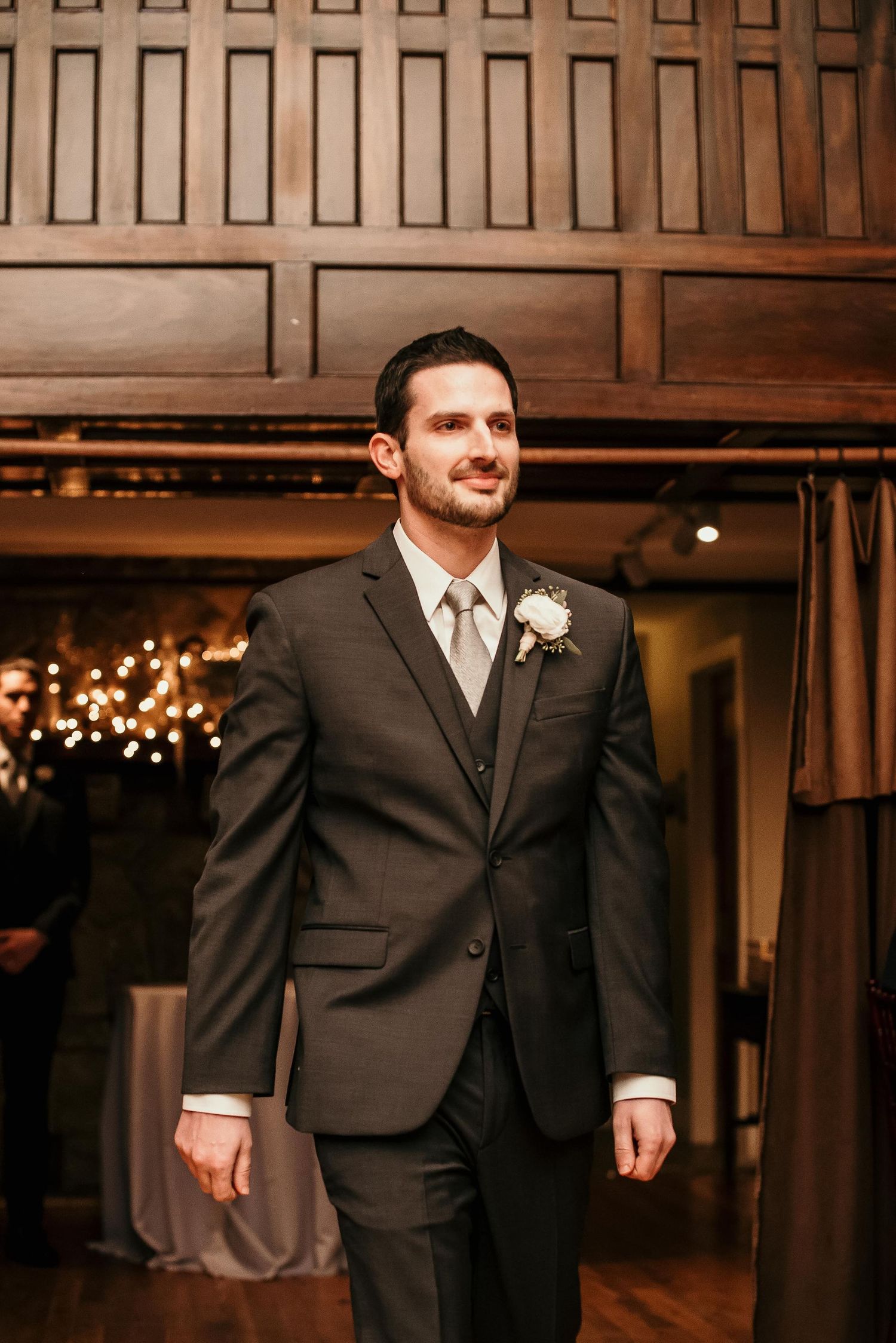 Groom in charcoal suit with white boutonniere smiling at wedding reception venue.
