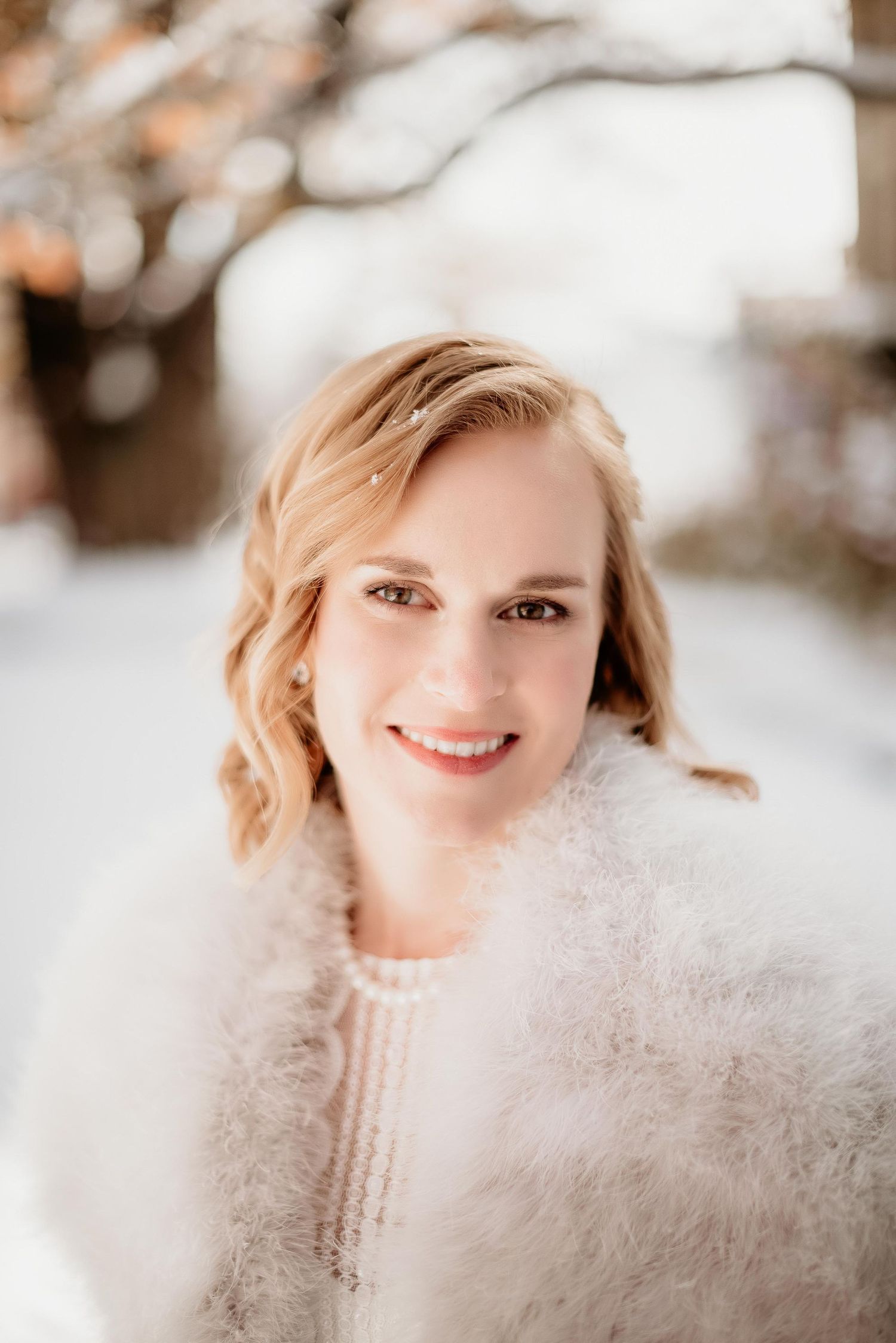 Smiling bride in white feather wrap and pearl necklace posing outdoors in a snowy winter setting.