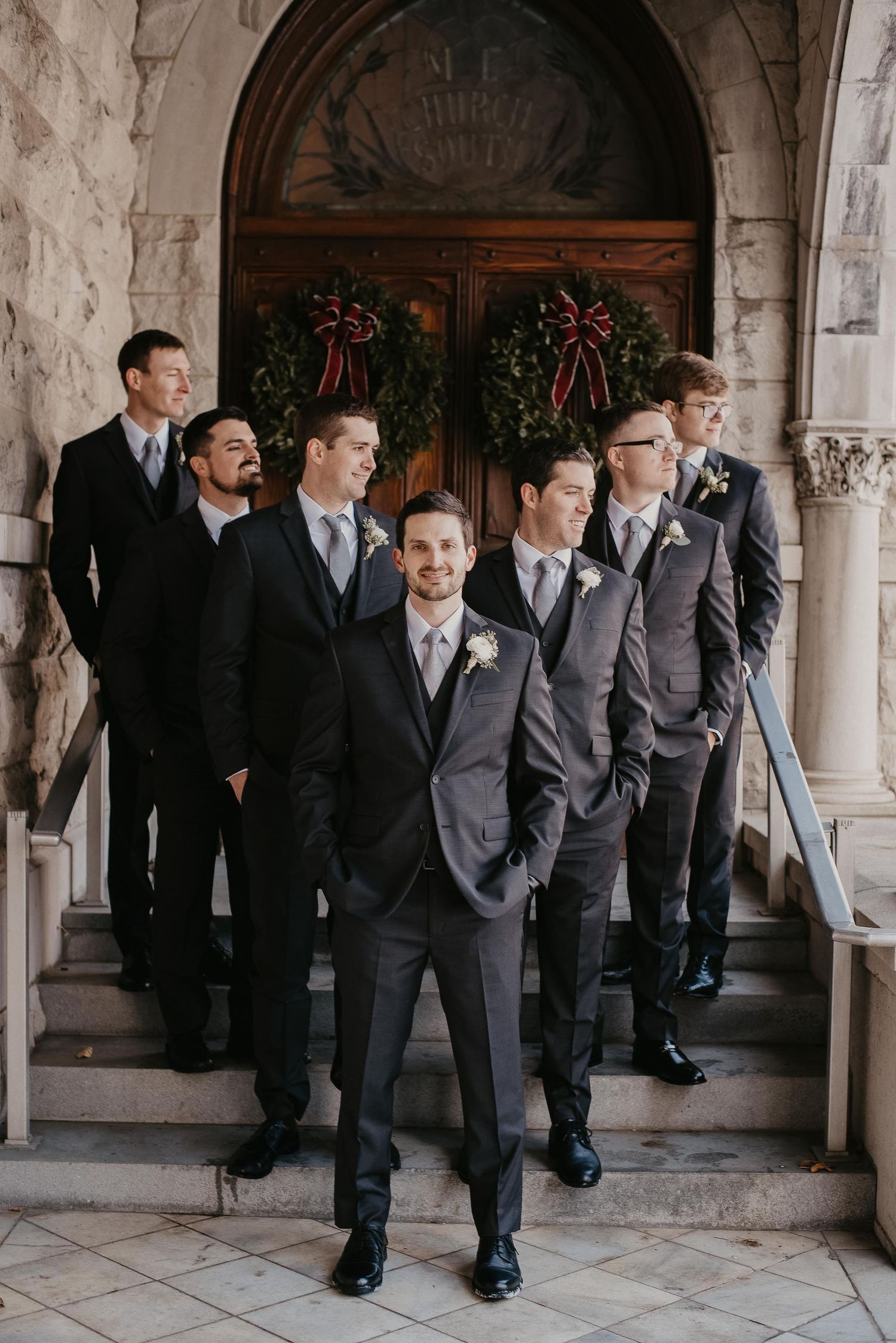 Groom and groomsmen in gray suits pose on church steps with holiday wreaths for a winter wedding.