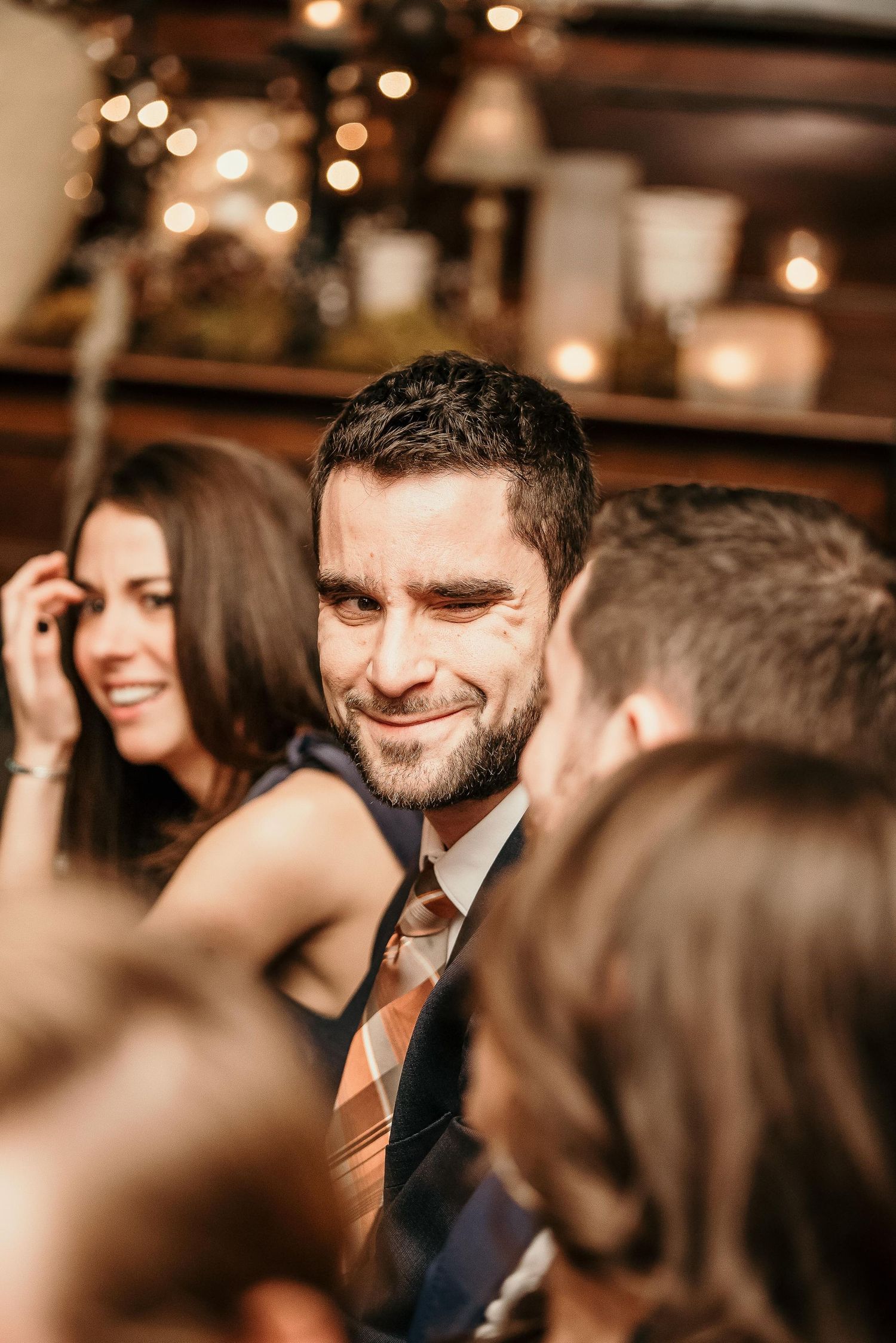 A smiling bearded man in a plaid tie mingles with guests at a warmly lit indoor social gathering or wedding reception.