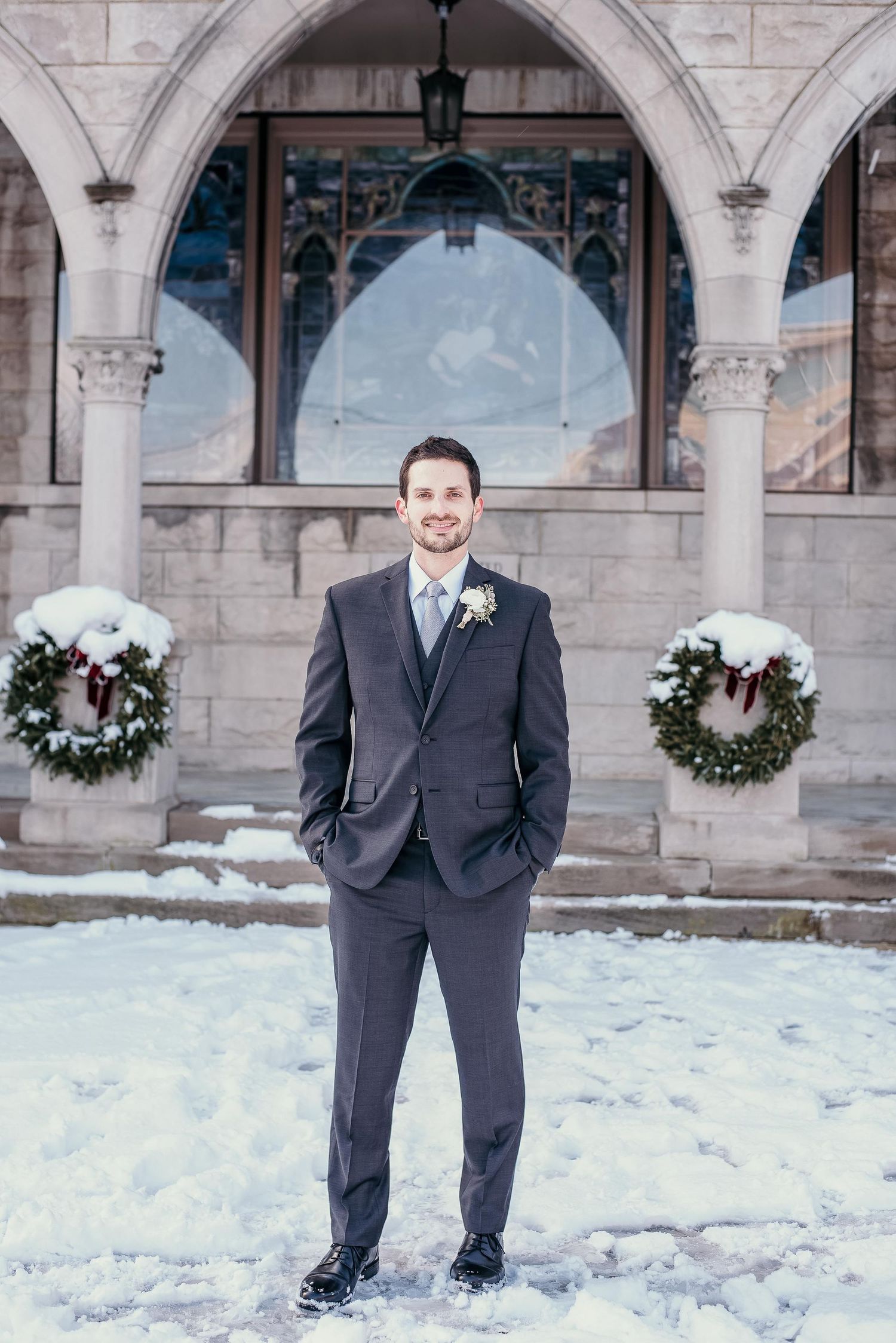 Groom in dark suit with boutonniere stands in snow outside stone church decorated with Christmas wreaths.