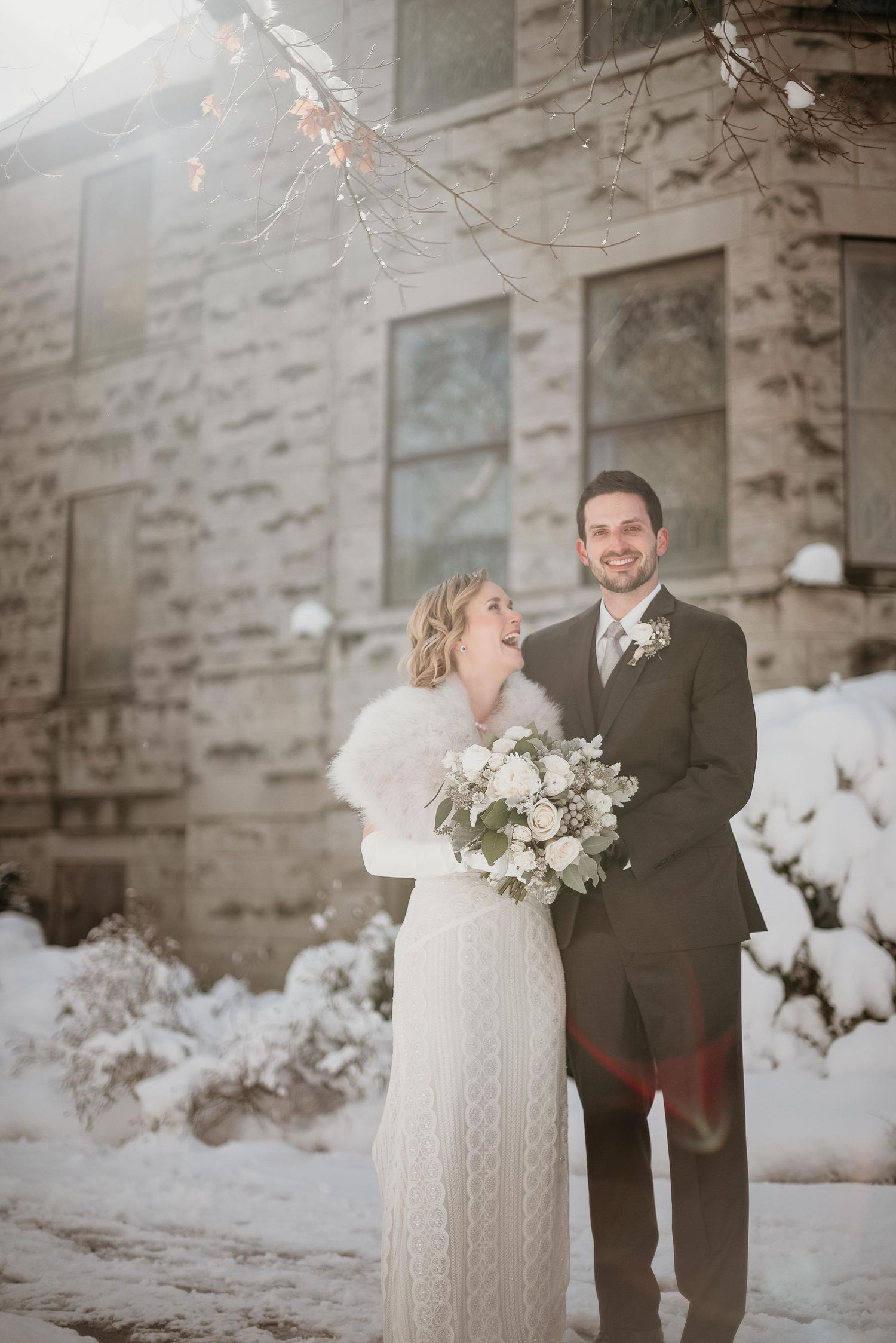 Bride and groom smiling together in snow outside stone building, bride holding white bouquet wearing fur wrap over lace gown.