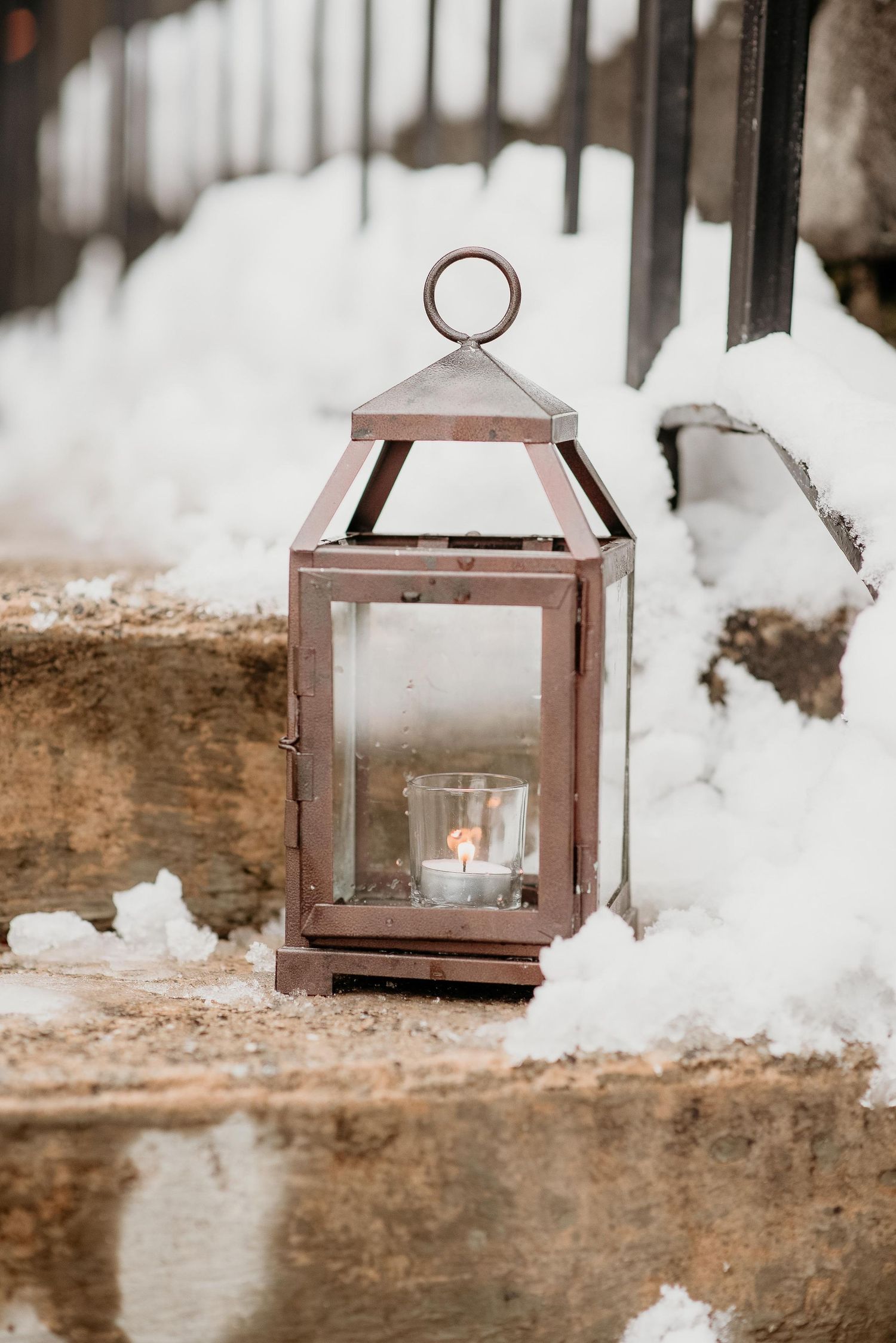 A rustic copper lantern with a lit candle sits on snowy stone steps in winter.