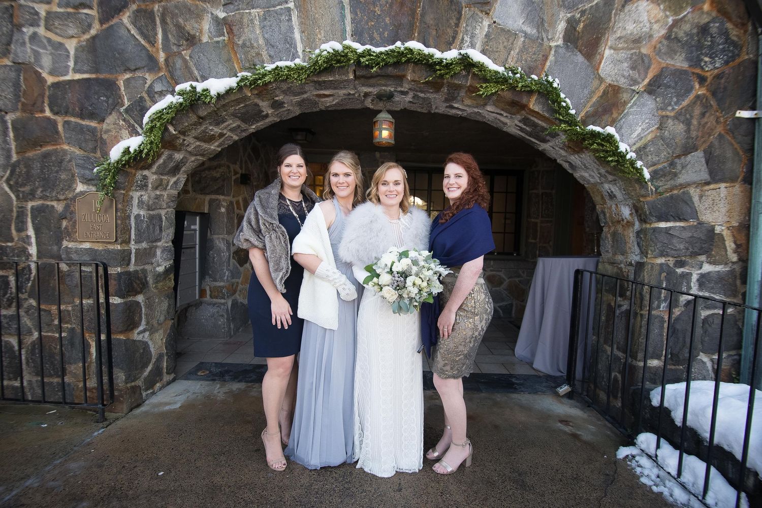 Bride in white gown poses with three bridesmaids in blue and gray dresses under a stone arch at a winter wedding.