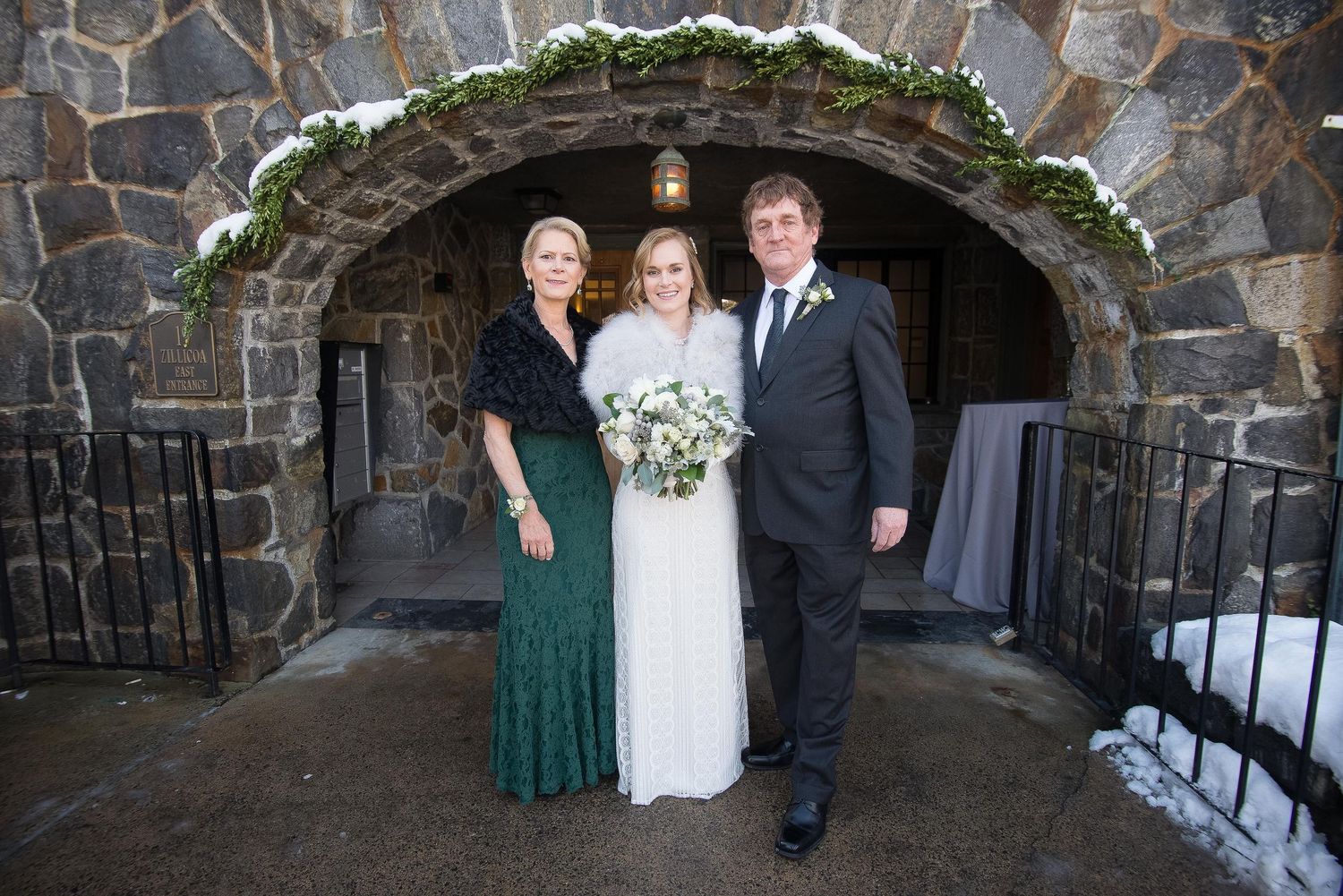 Bride in white gown poses with two guests outside a stone arch venue dusted with snow at a winter wedding.