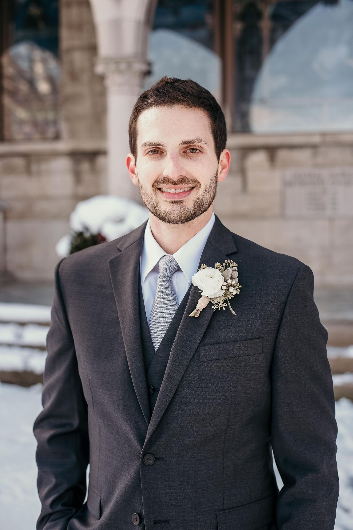 Smiling groom in dark suit with white boutonniere poses outdoors in winter setting near stone building.