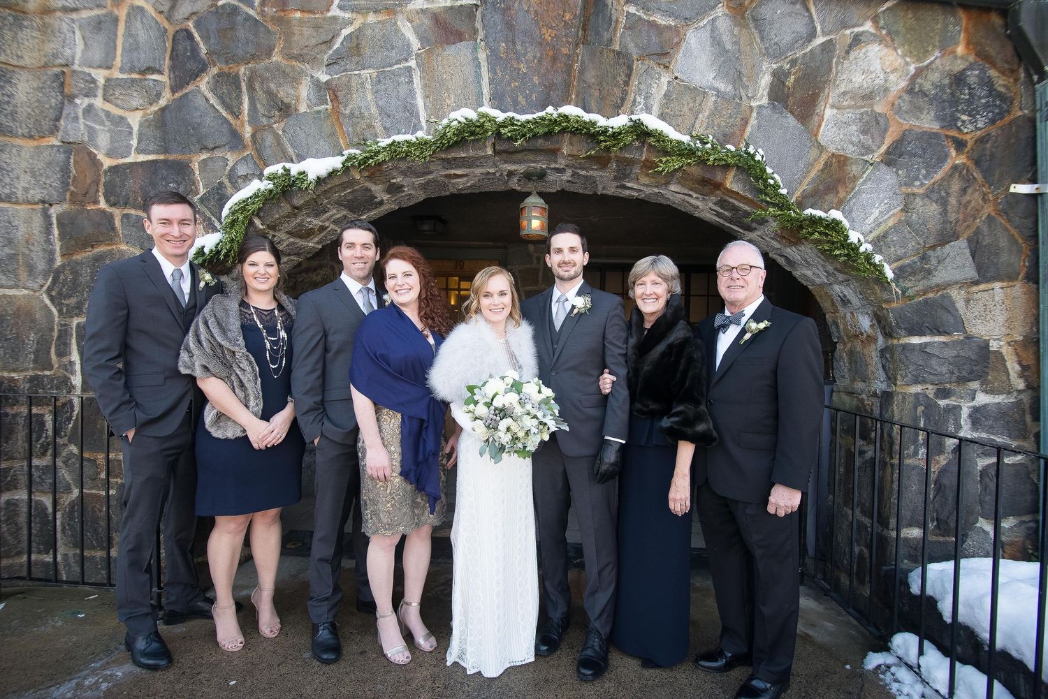 Wedding party poses in front of stone archway in winter, bride holding white bouquet with family and groomsmen.