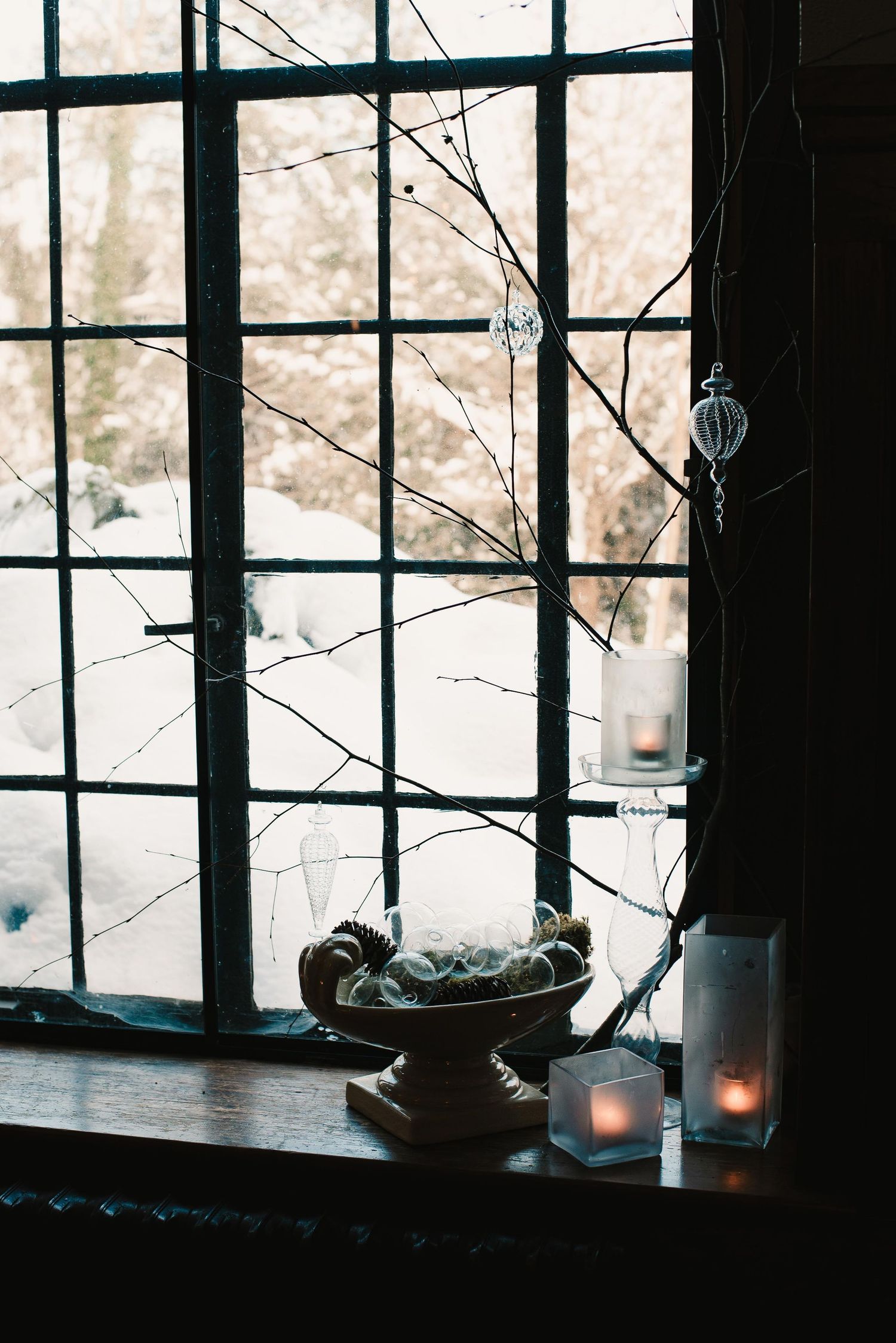 Cozy winter window display with bare branches, glass ornaments, candles, and a decorative bowl on a wooden ledge.