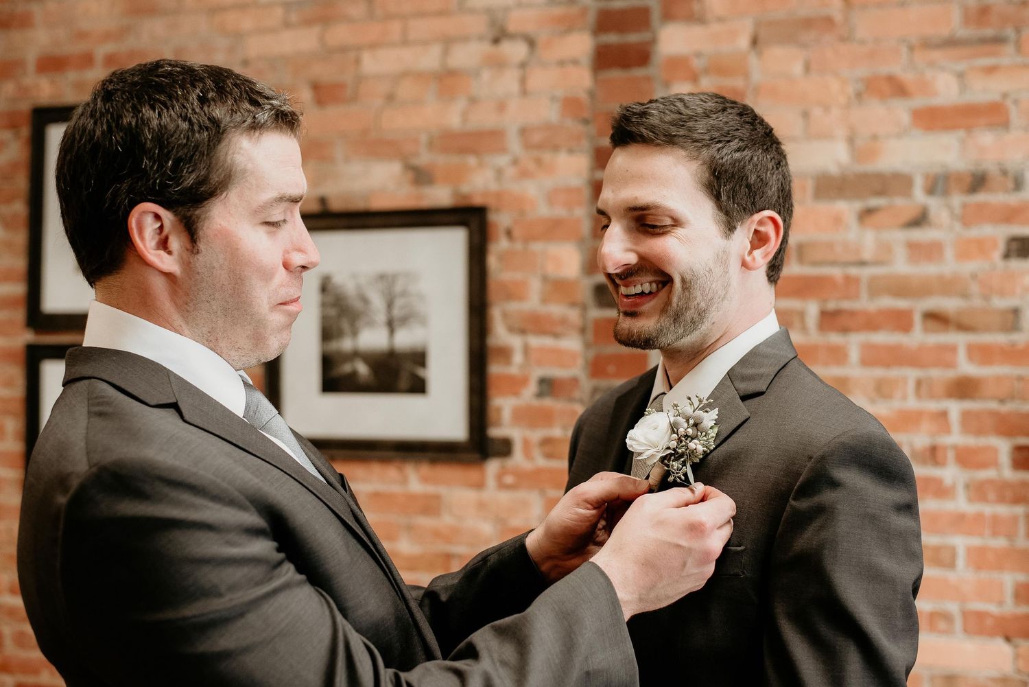 Groomsman pinning a white boutonniere on the groom's dark suit lapel, smiling, with a brick wall backdrop.