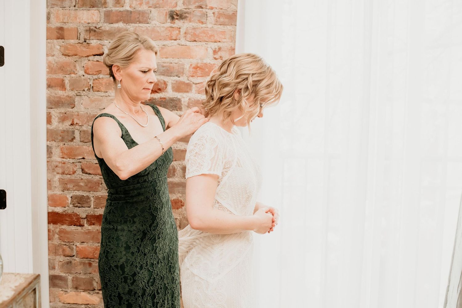 Mother helping bride into white lace wedding dress near a sunlit window and exposed brick wall.