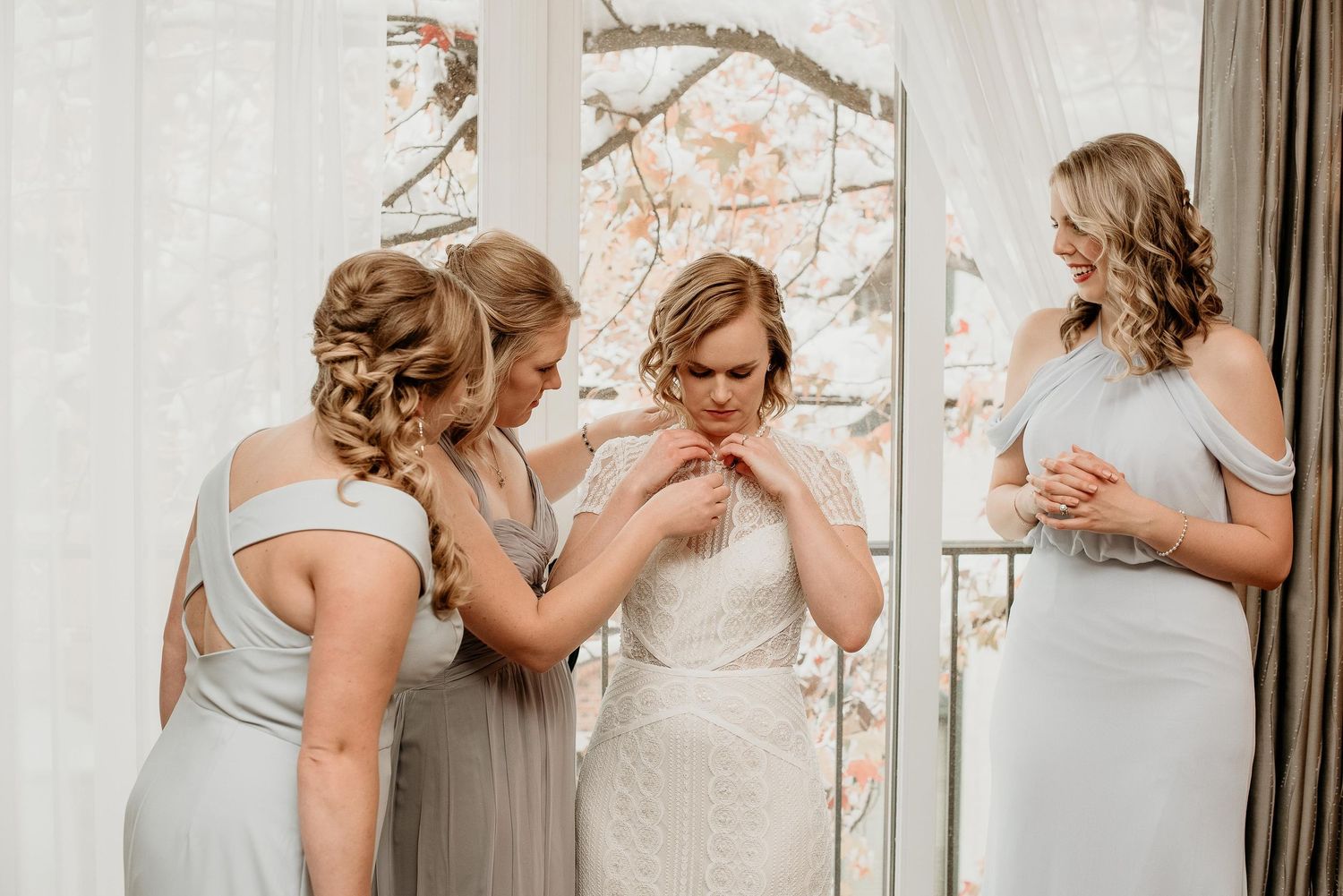 Bridesmaids in silver gowns help bride fasten her white lace wedding dress near a sunlit window.