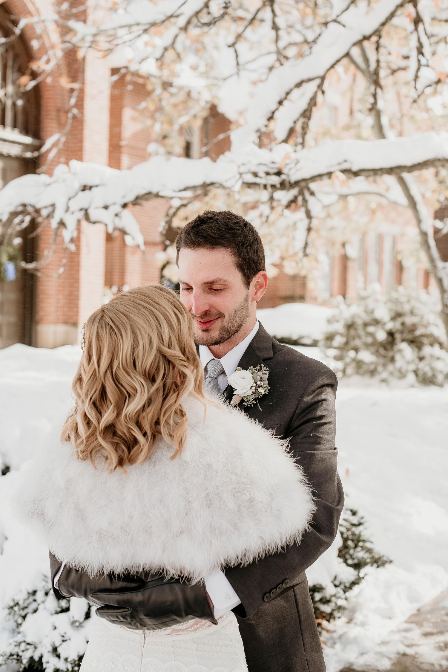 Bride in white fur wrap and groom in gray suit share a romantic moment outdoors in a snowy winter wedding setting.